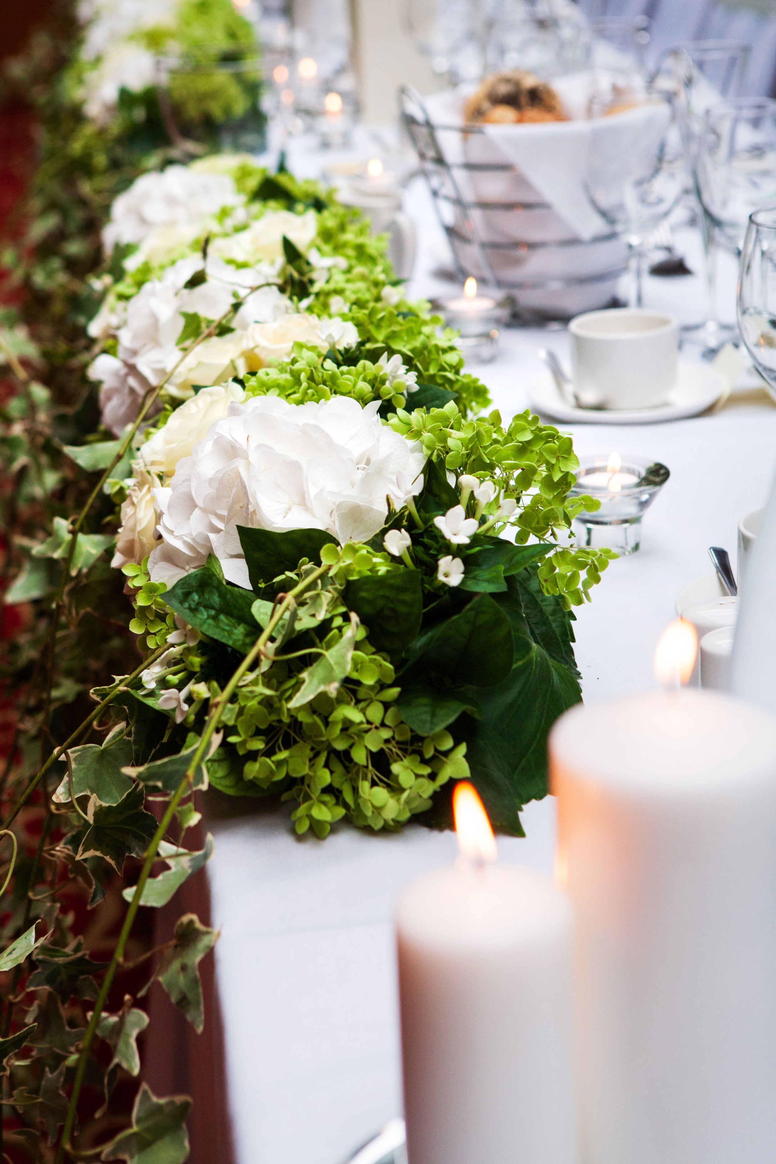 A close-up of a table decorated with a floral centerpiece of white and green flowers, surrounded by lit candles at an elegant event or gathering.