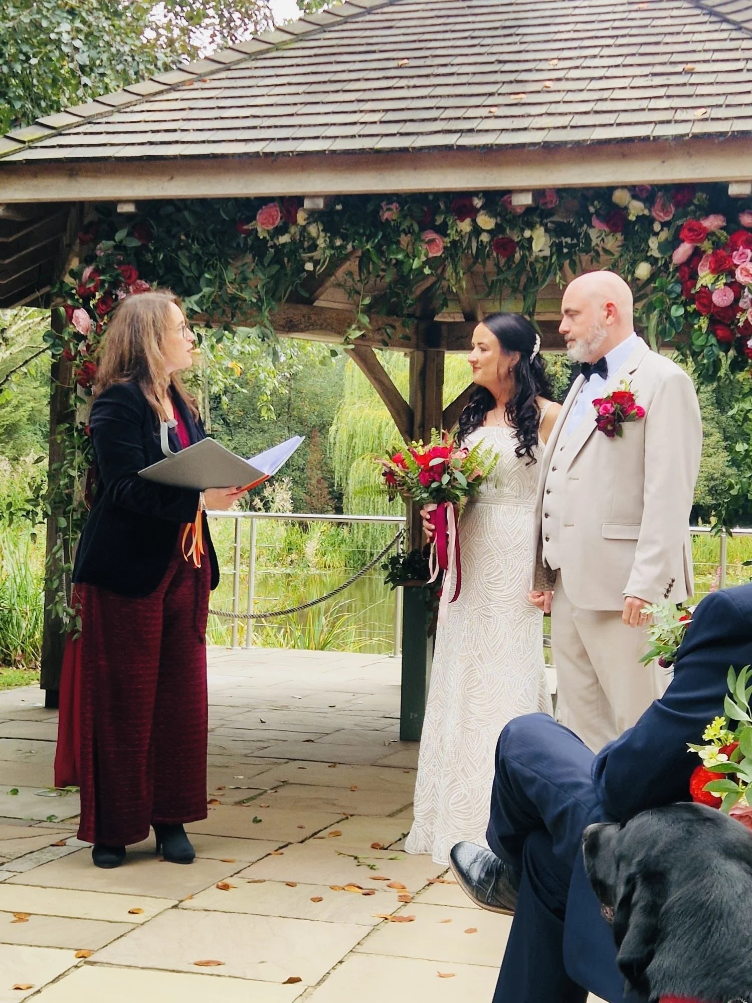 A wedding ceremony outdoors with a bride and groom facing an officiant under a wooden gazebo decorated with red and pink roses. The bride is holding a bouquet of red flowers, and the groom is wearing a light-colored suit with a red boutonniere.