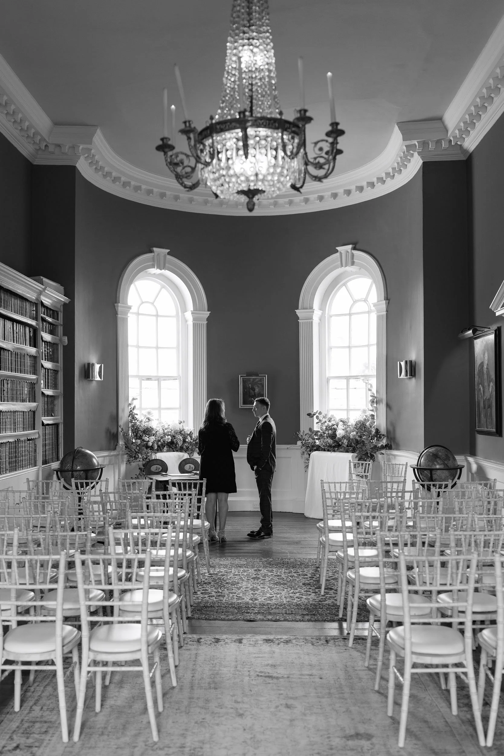 A black and white photo of a room with high ceilings, large arched windows, and elegant decorations. Two people are standing in the centre engaged in conversation.