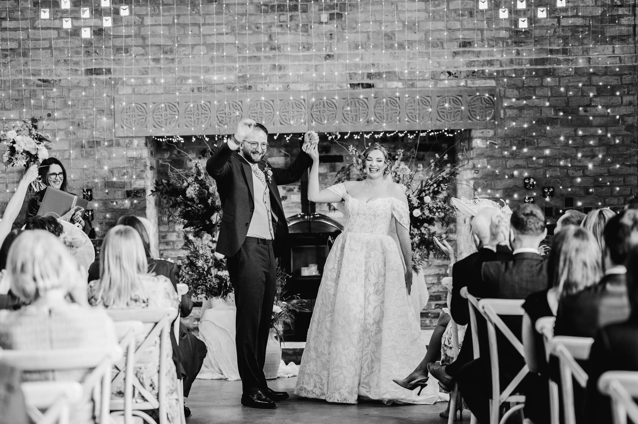 Black and white photo of a wedding reception with a couple dancing, the groom is holding the bride's hand in a celebratory gesture, surrounded by seated guests in a decorated venue with string lights and floral arrangements.