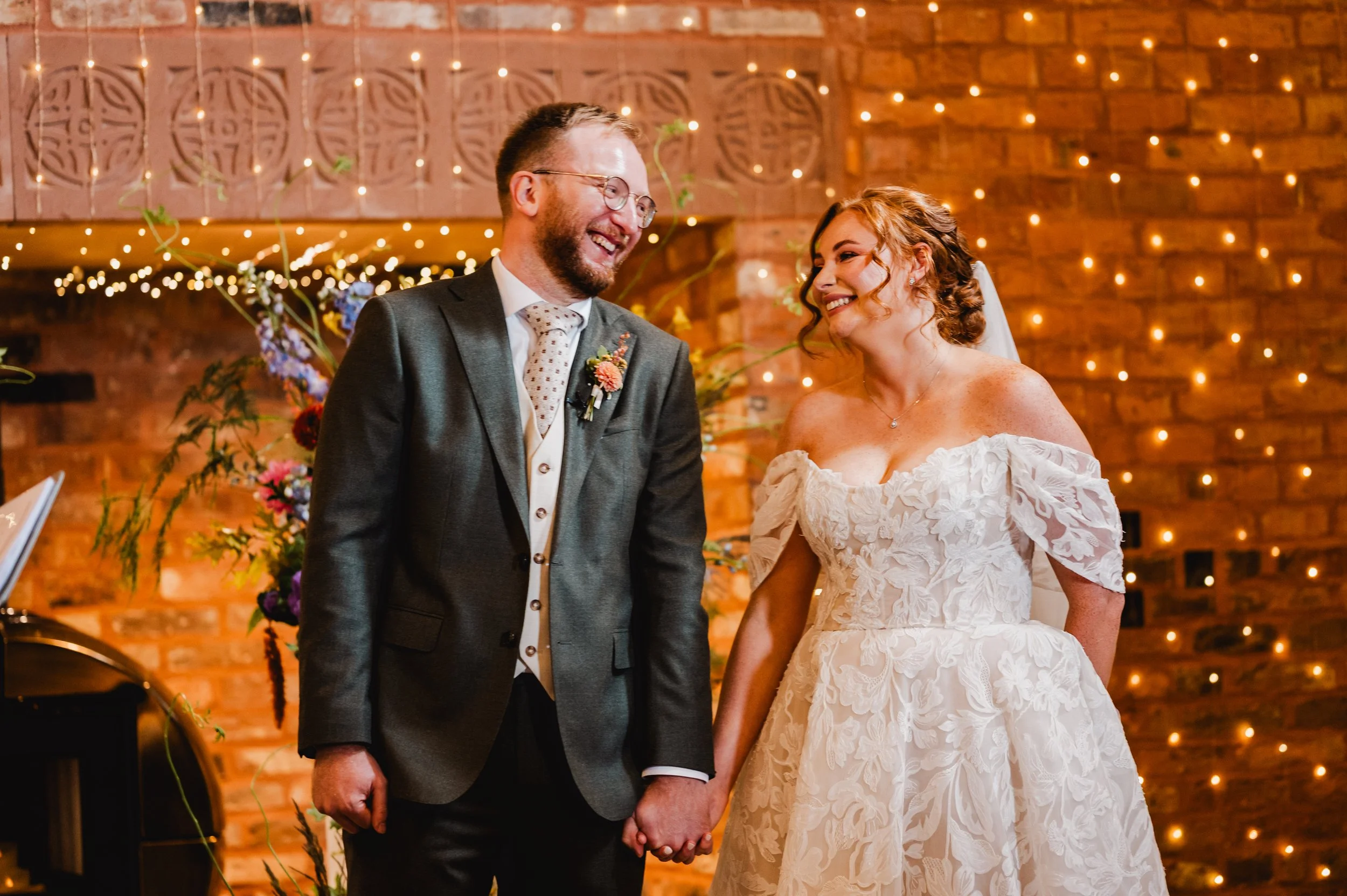 A bride and groom holding hands during their wedding ceremony, smiling at each other against a brick wall decorated with string lights and floral arrangements.