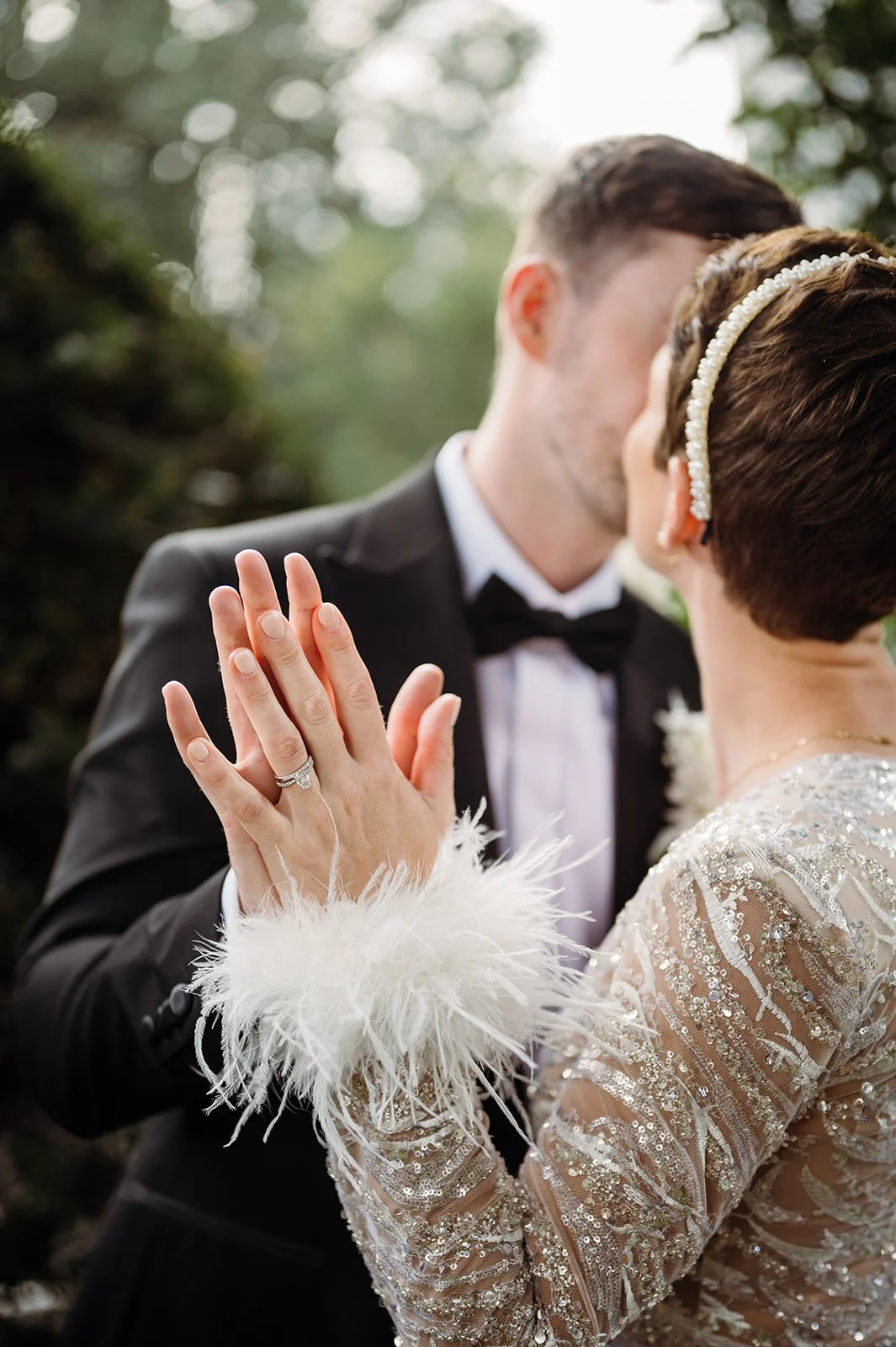 A couple dressed in wedding attire, kissing outdoors. The woman is showing her wedding ring with her hand raised, wearing a glamorous jumpsuit with sequins and feathers. The man is wearing a black tuxedo with a bow tie, with soft natural background.