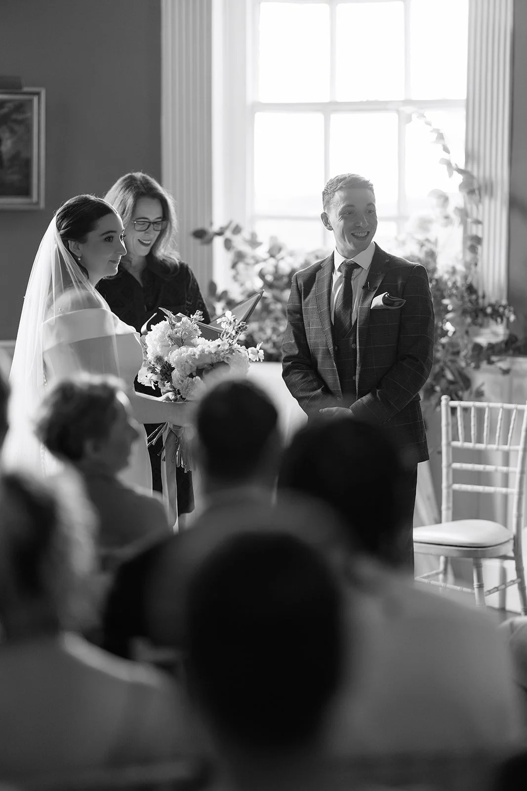 A wedding ceremony with a bride, a groom, and an officiant standing before seated guests, illuminated by natural light streaming through a large window.