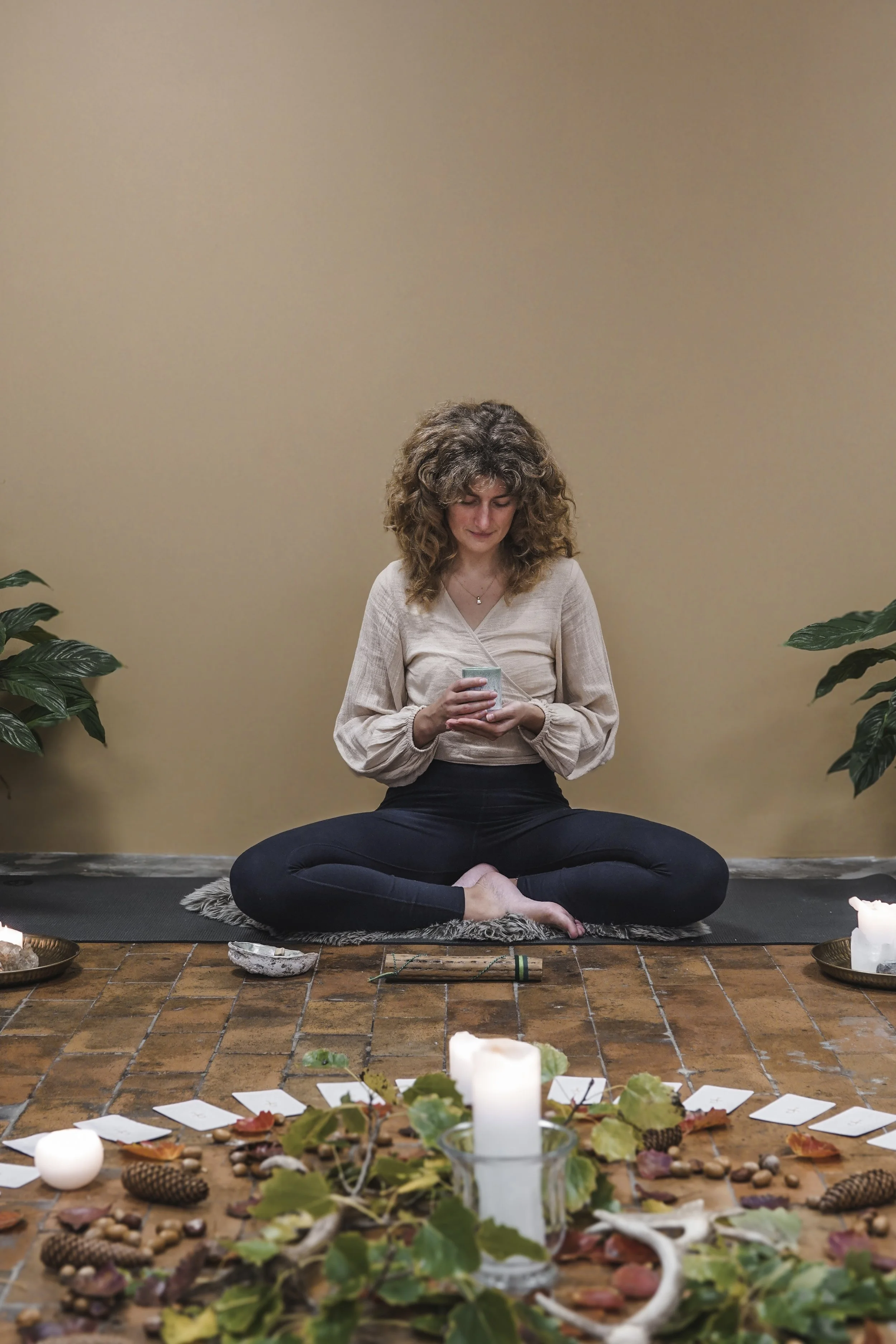 En kvinde sidder krydsbenet på en yogamåtte foran en lavtbord med lysende stearinlys, blade, nødder, pinde og kort, omgivet af grønne planter, mens hun holder en kop.