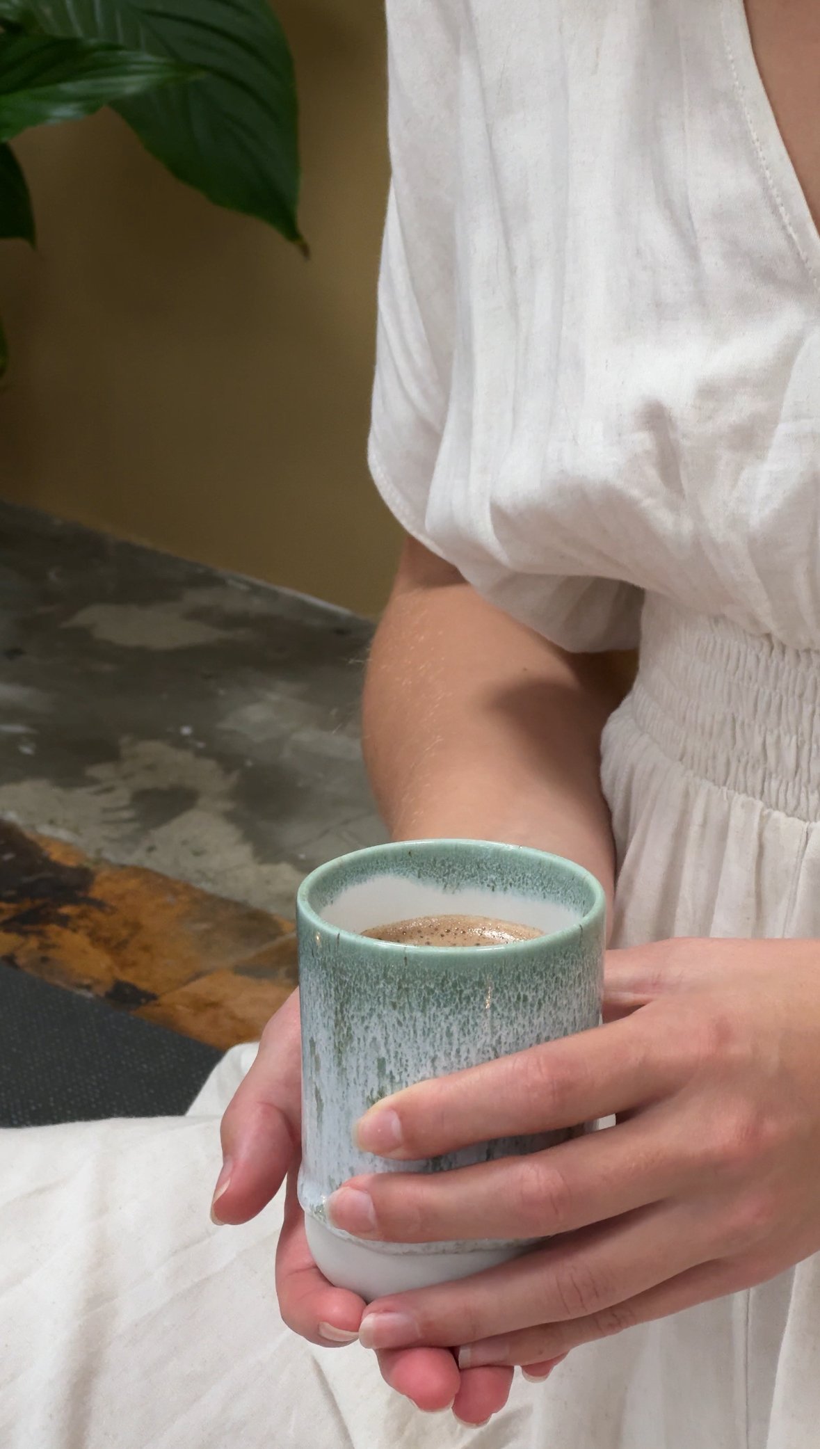En person holder en kop varm chokolade eller kaffe, iført en hvid kjole eller trøje, med en grøn plante på baggrund.