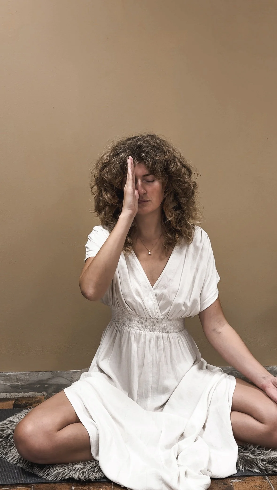 Kvindelig person sidder i korslagte ben på gulvet med øjnene lukkede og en hånd mod panden, iført en hvid kjole, foran en beige væg.