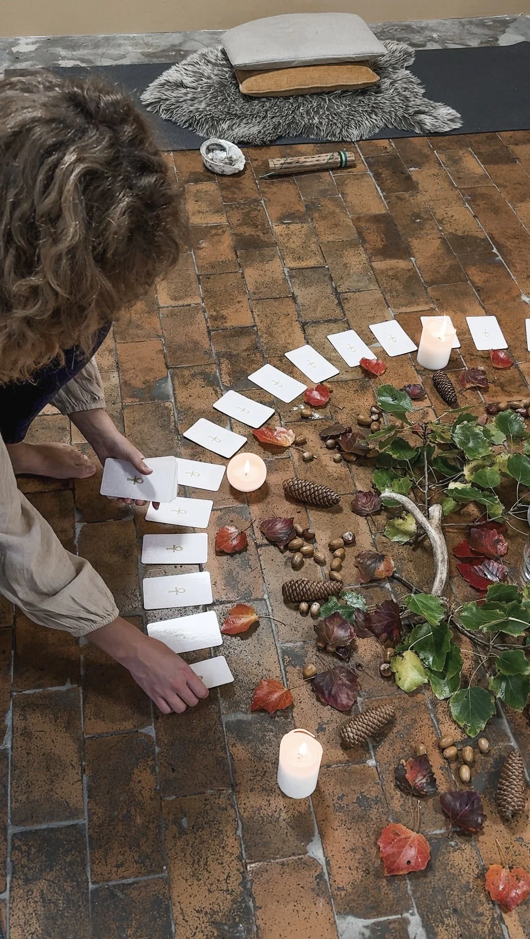 Person, der arrangerer kort i en cirkel omgivet af efterårsblade, kogler og nødder, med stearinlys på en stenflisegulv.