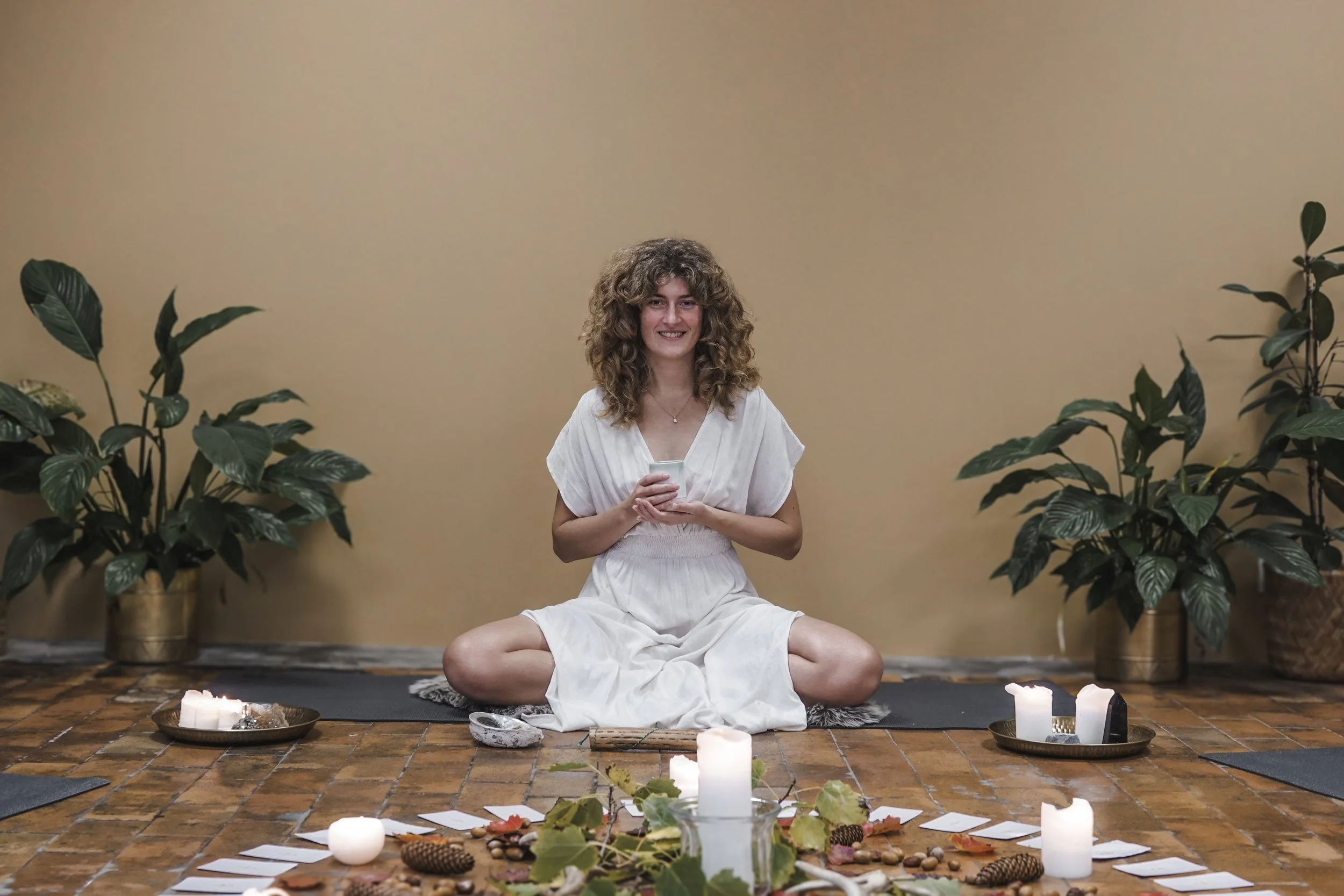 En kvinde med krøllet hår, iført hvidt kjole, sidder krydsbenet på gulvet og holder en kop, omgivet af lys, blade og naturmaterialer, i et rum med beige væg og grønne planter.