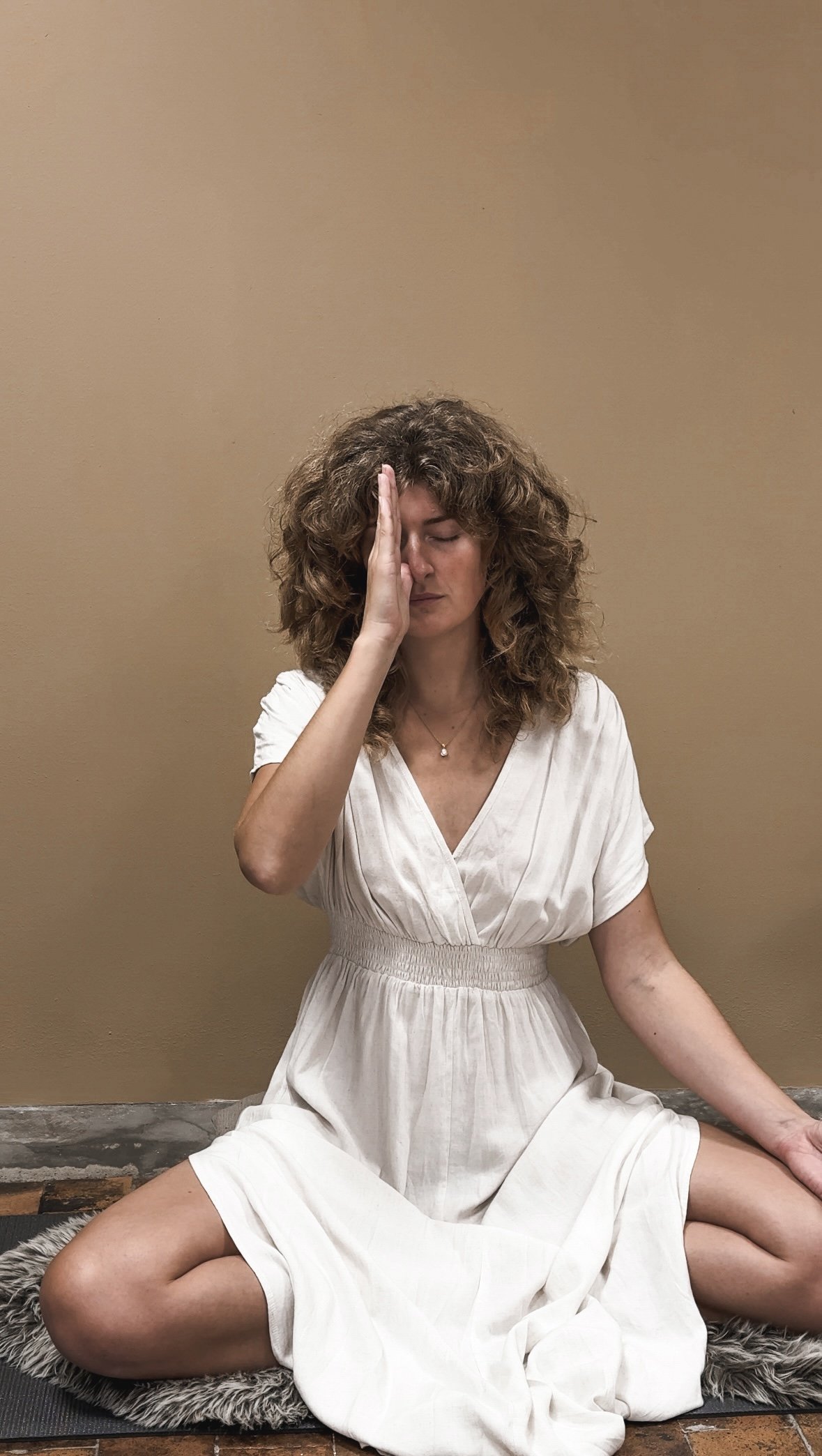 Kvinde sidder på knæ med lukkede øjne, hånd på panden, foran en beige væg, klædt i hvid kjole, på en grågette med pelskant.