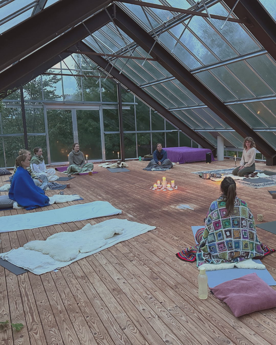 En gruppe mennesker sidder på gulvet i et lyst trækonstruktion rum med glasvægge, og deltager i en meditations- eller yogaøvelse, omgivet af lejlighedsvis lysende stearinlys og tæpper.