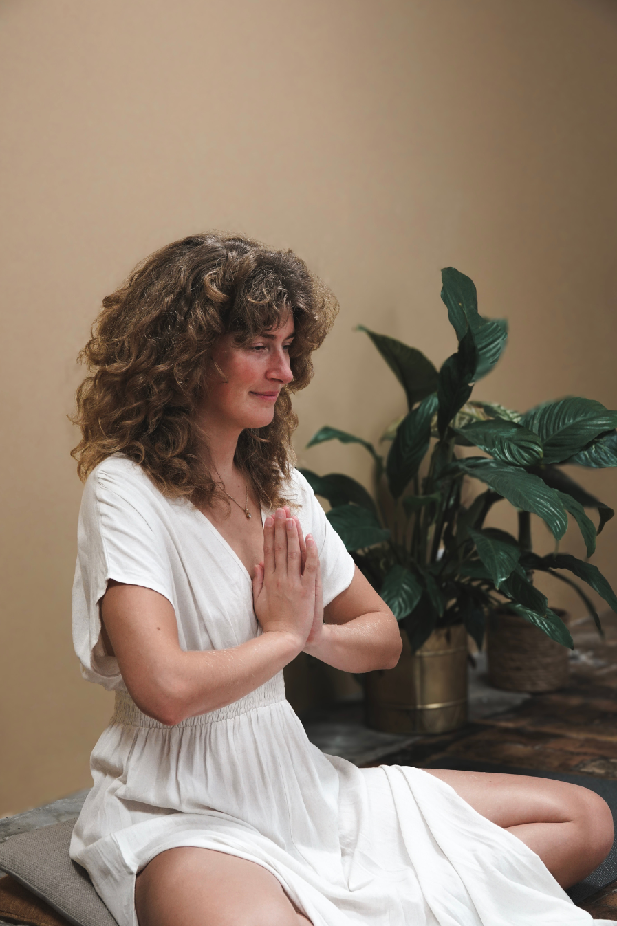 Kvinde med krøllet hår sidder på gulvet med håndflader together foran brystet, praktiserer meditation eller yoga, foran et grønt bladplanter.