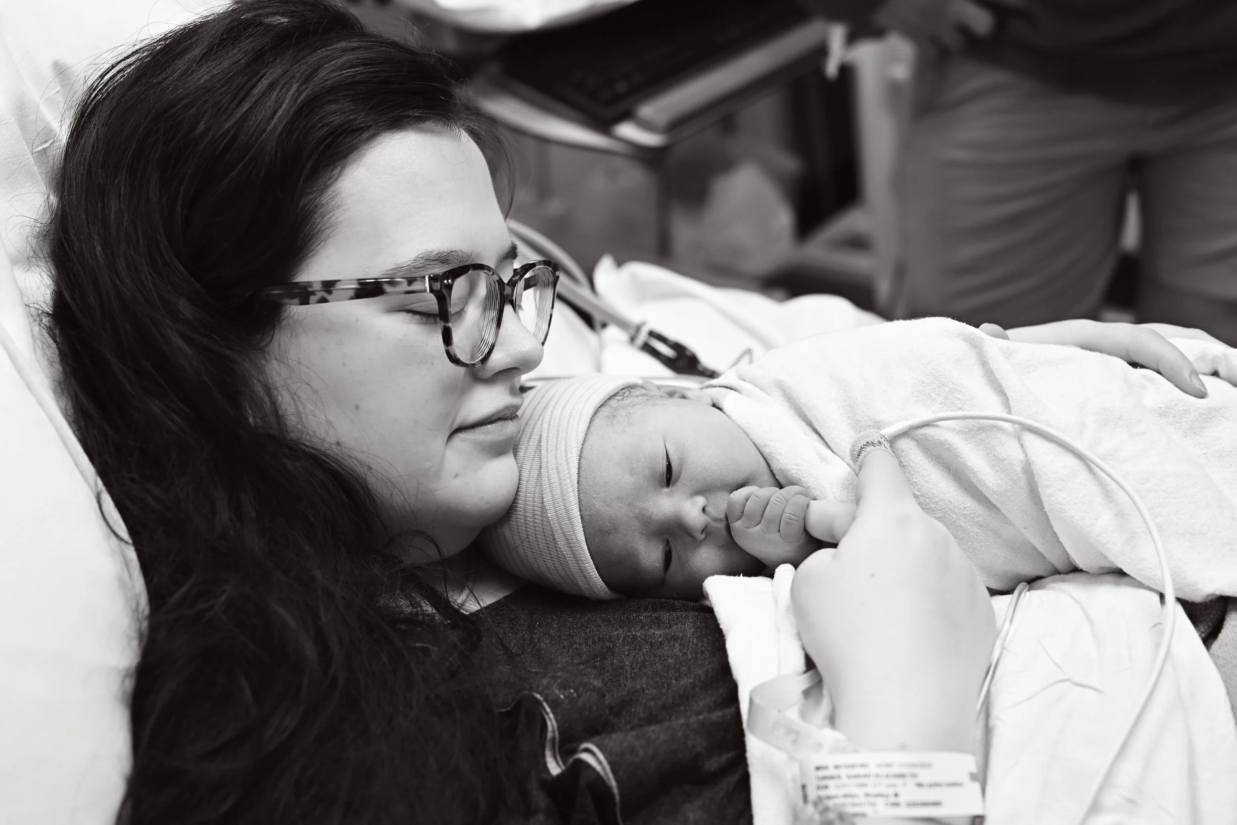 A woman with glasses holding a newborn baby in a hospital bed.