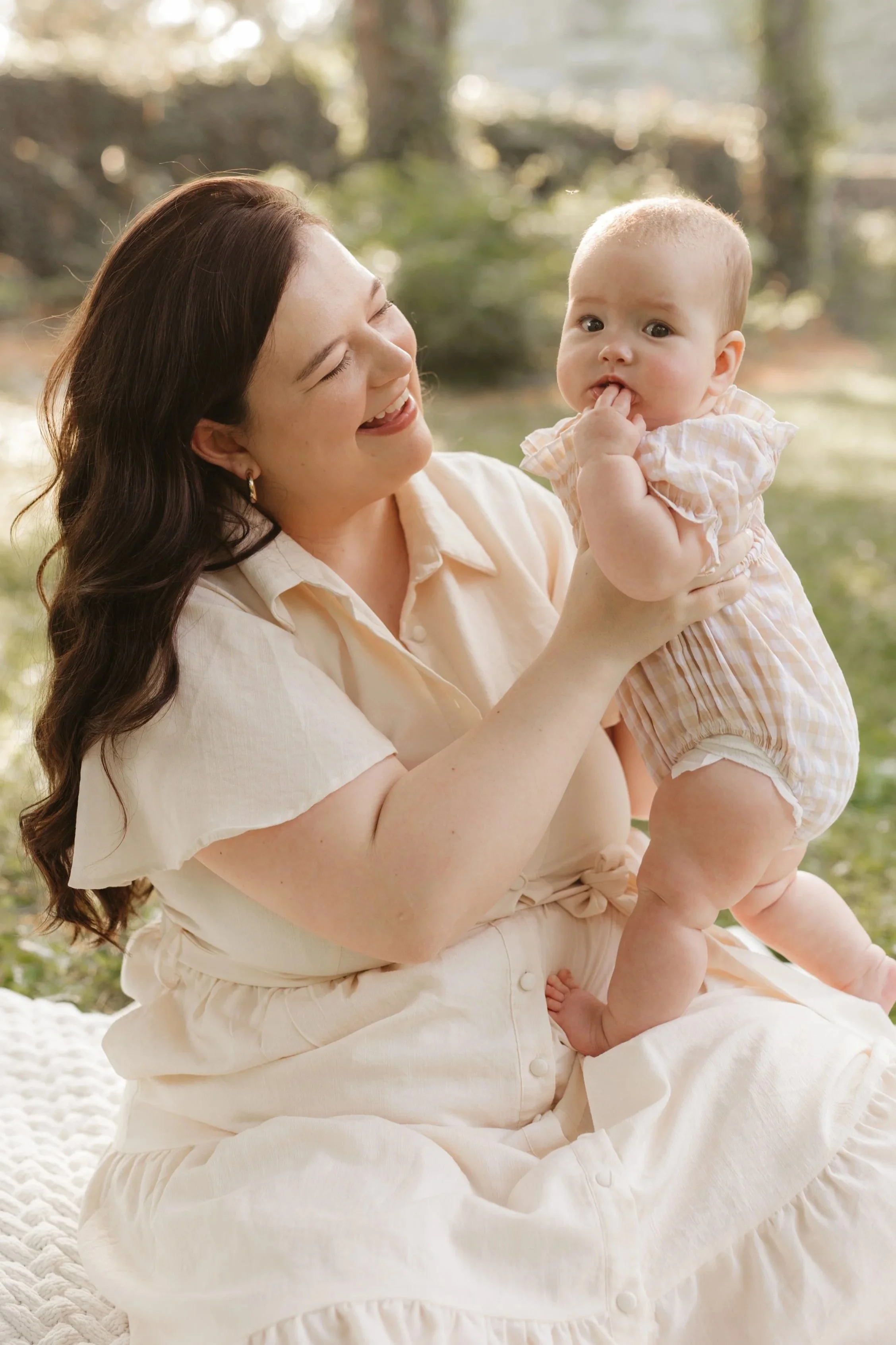 A woman with long brown hair, dressed in a light beige dress, is smiling and holding a baby girl in her arms. The baby is wearing a light pink plaid romper and has her finger in her mouth. They are outdoors in a natural setting with trees and greenery in the background, enjoying a sunny day.