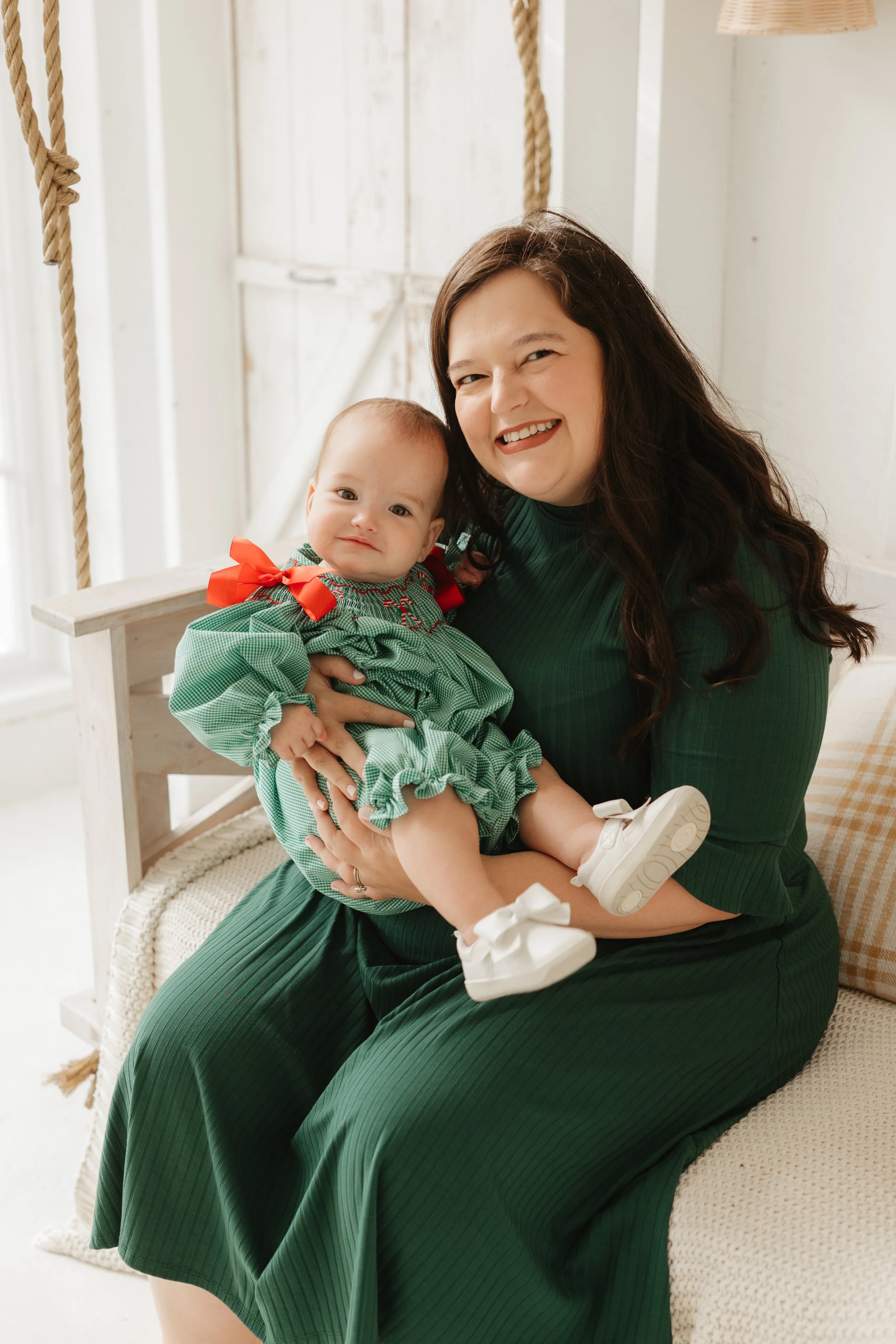 A woman in a green dress holding a baby girl in a green dress with red bow, sitting on a cream-colored couch in a bright room.