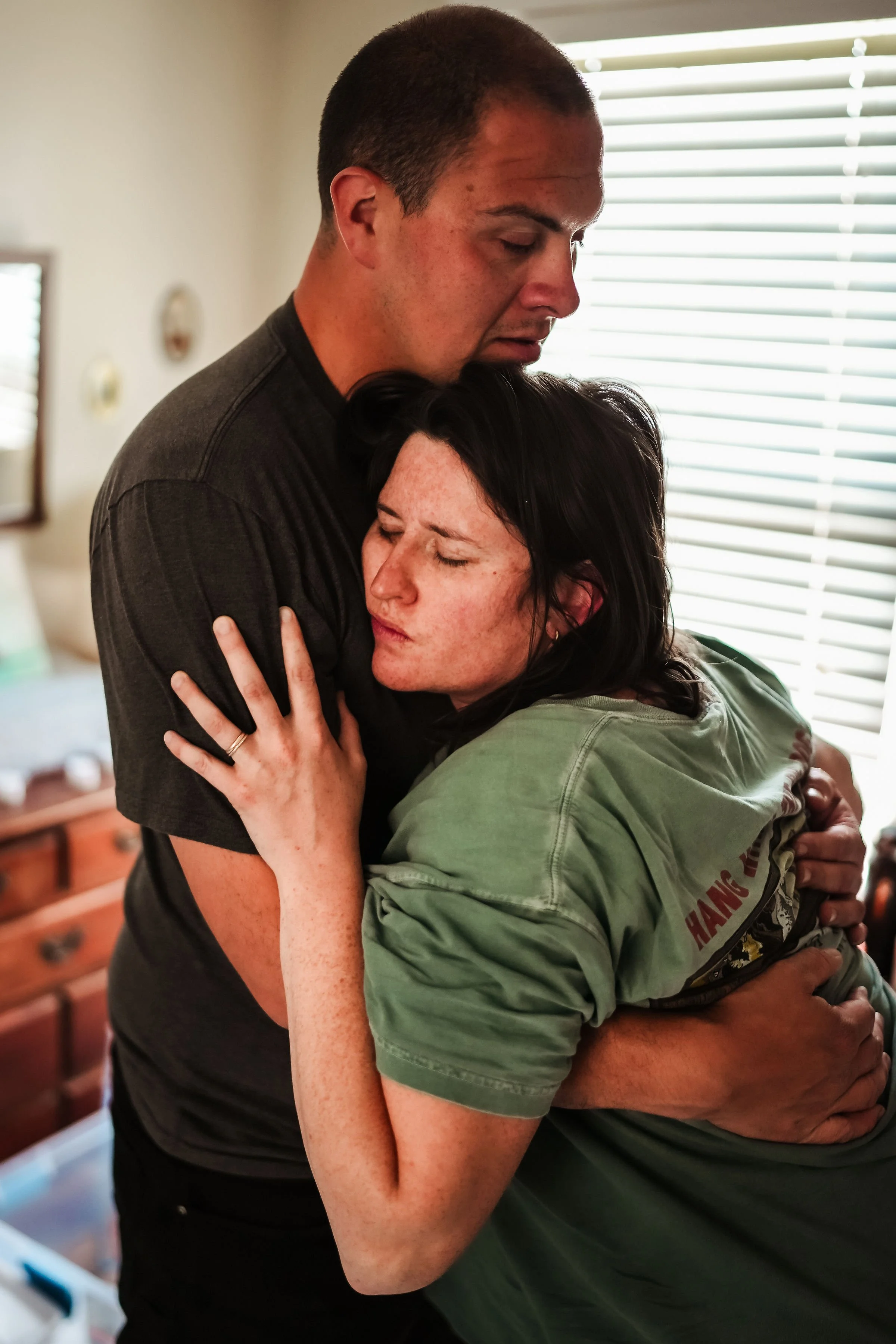 A man and a woman sharing a heartfelt hug, eyes closed, in a cozy room with blinds covering the window.