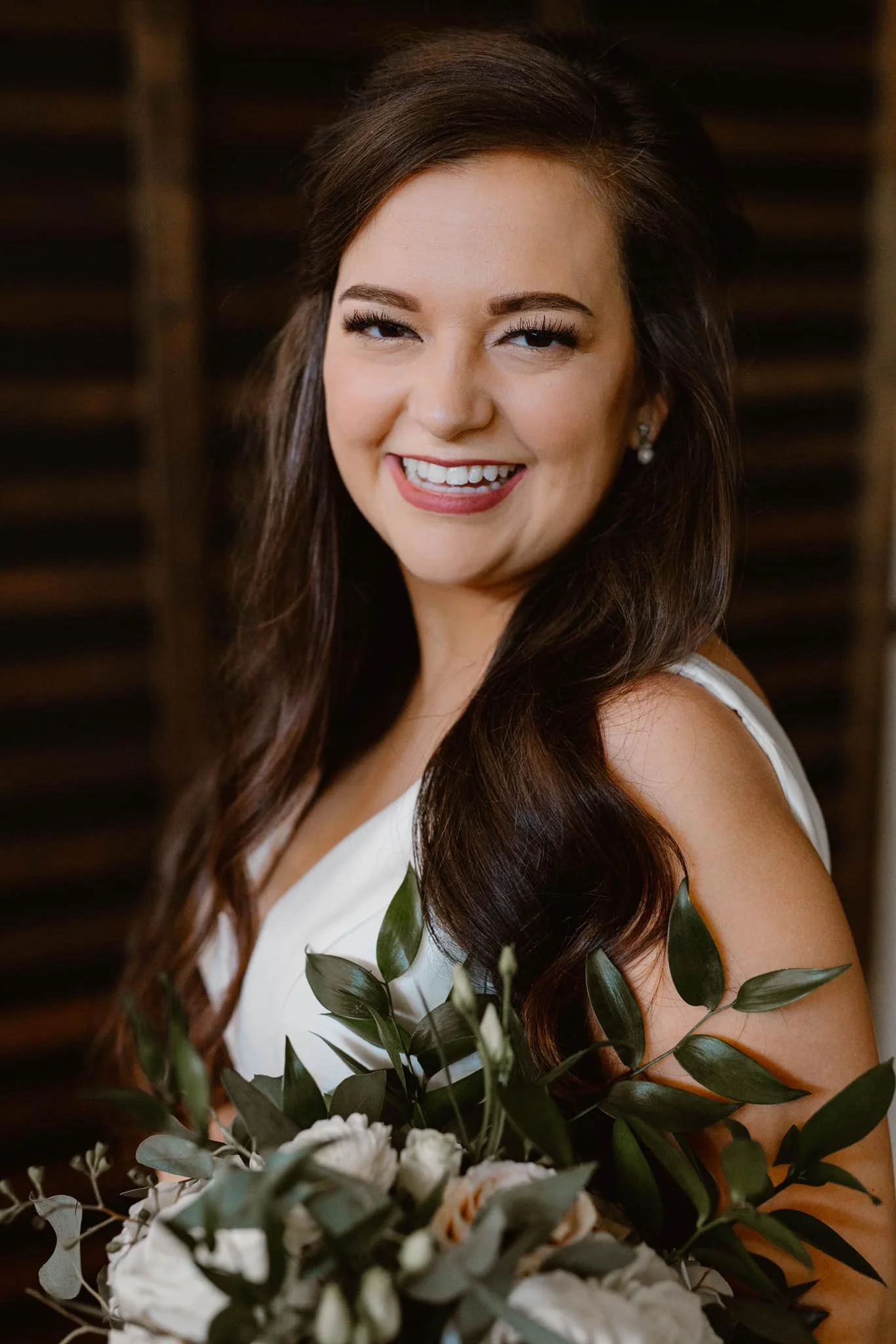 A woman smiling, holding a bouquet of white roses and green leaves, wearing a white dress with dark brown hair