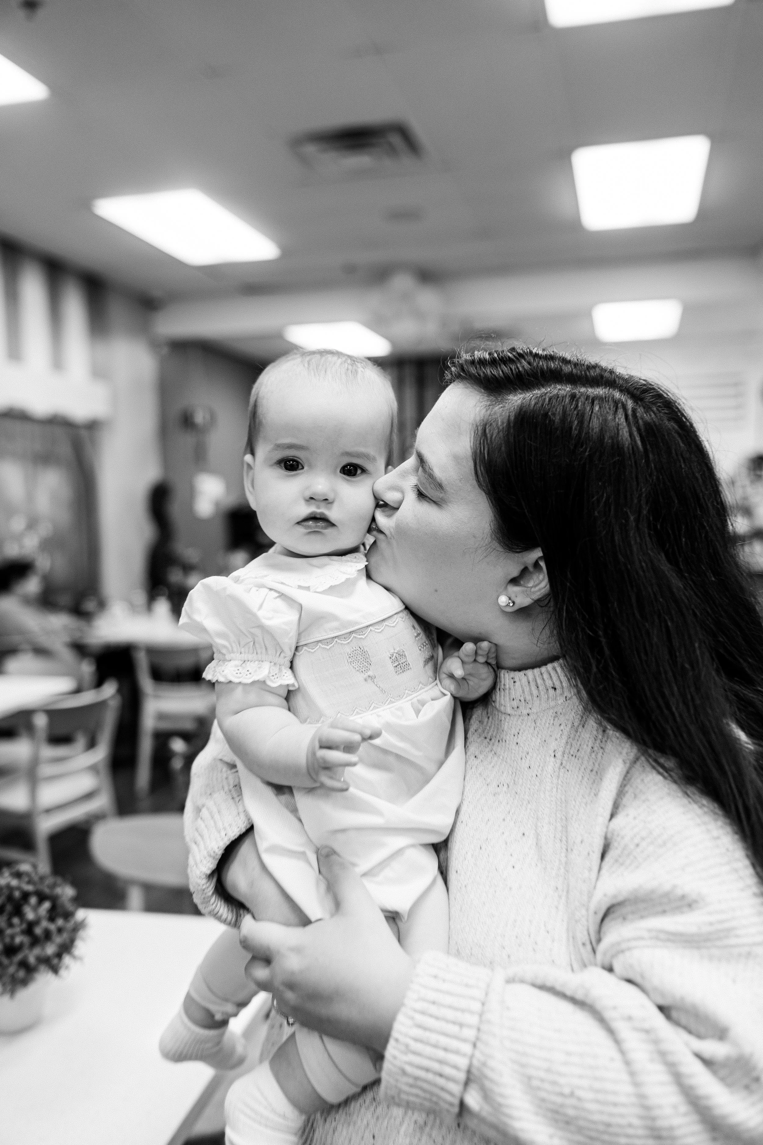 A woman holding and kissing a baby girl, in an indoor setting with tables and chairs.