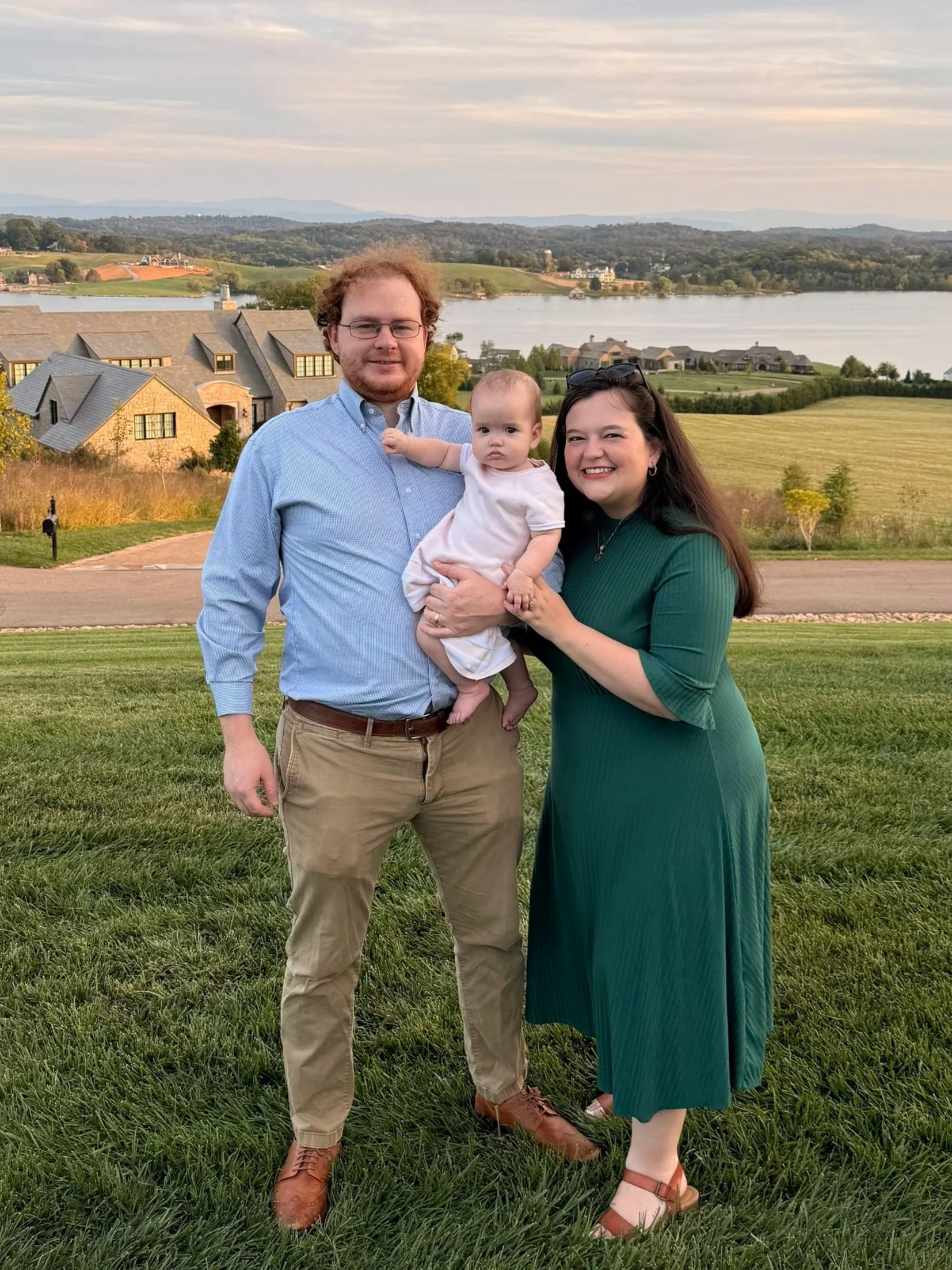 A family of three standing on a grassy area with a scenic lake and hills in the background during sunset. The man is holding a young child, and the woman is smiling, wearing a green dress.