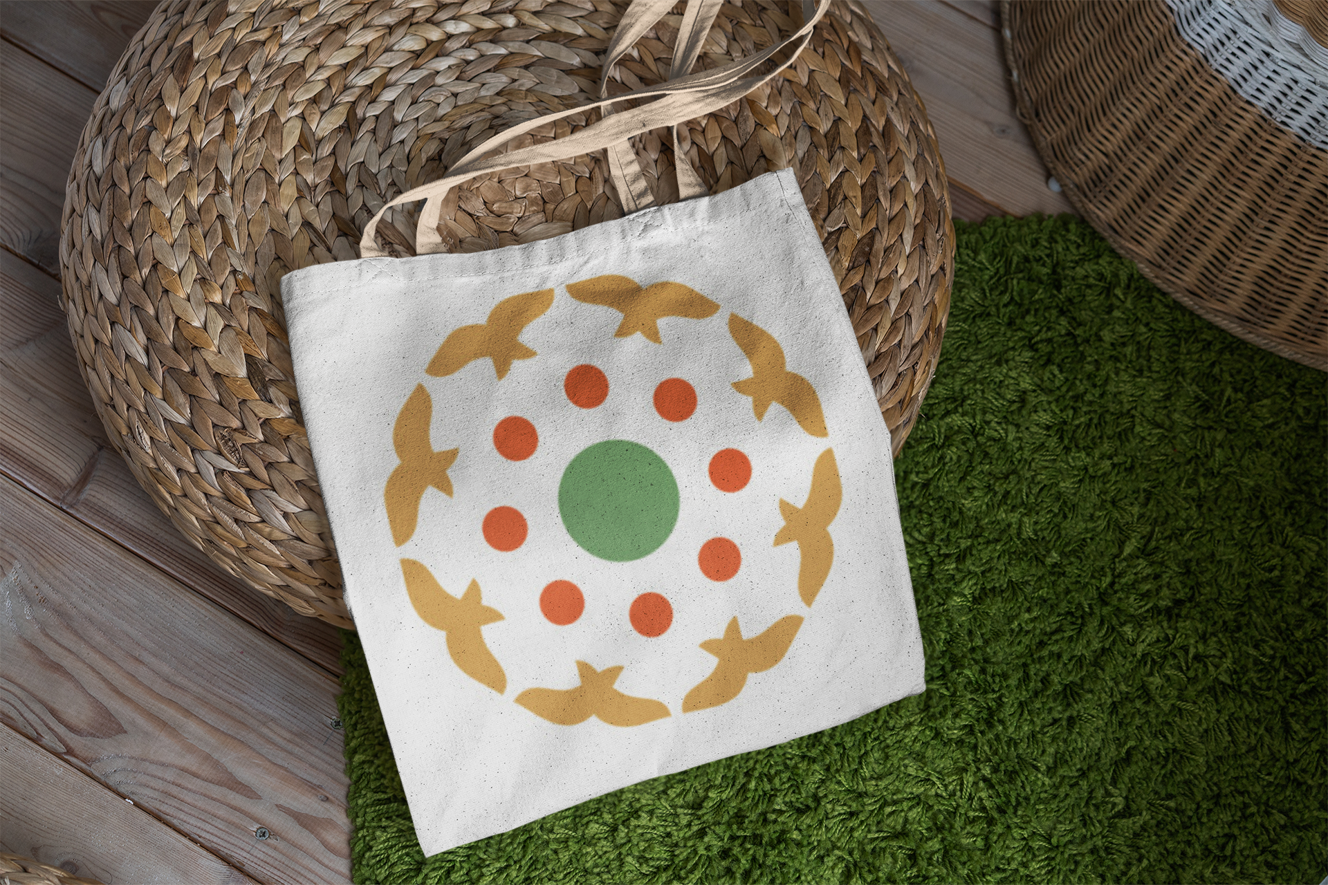A white tote bag with a printed circular pattern of yellow birds, orange dots, and a green center, resting on a round woven basket placed on a green textured rug and wooden floor.