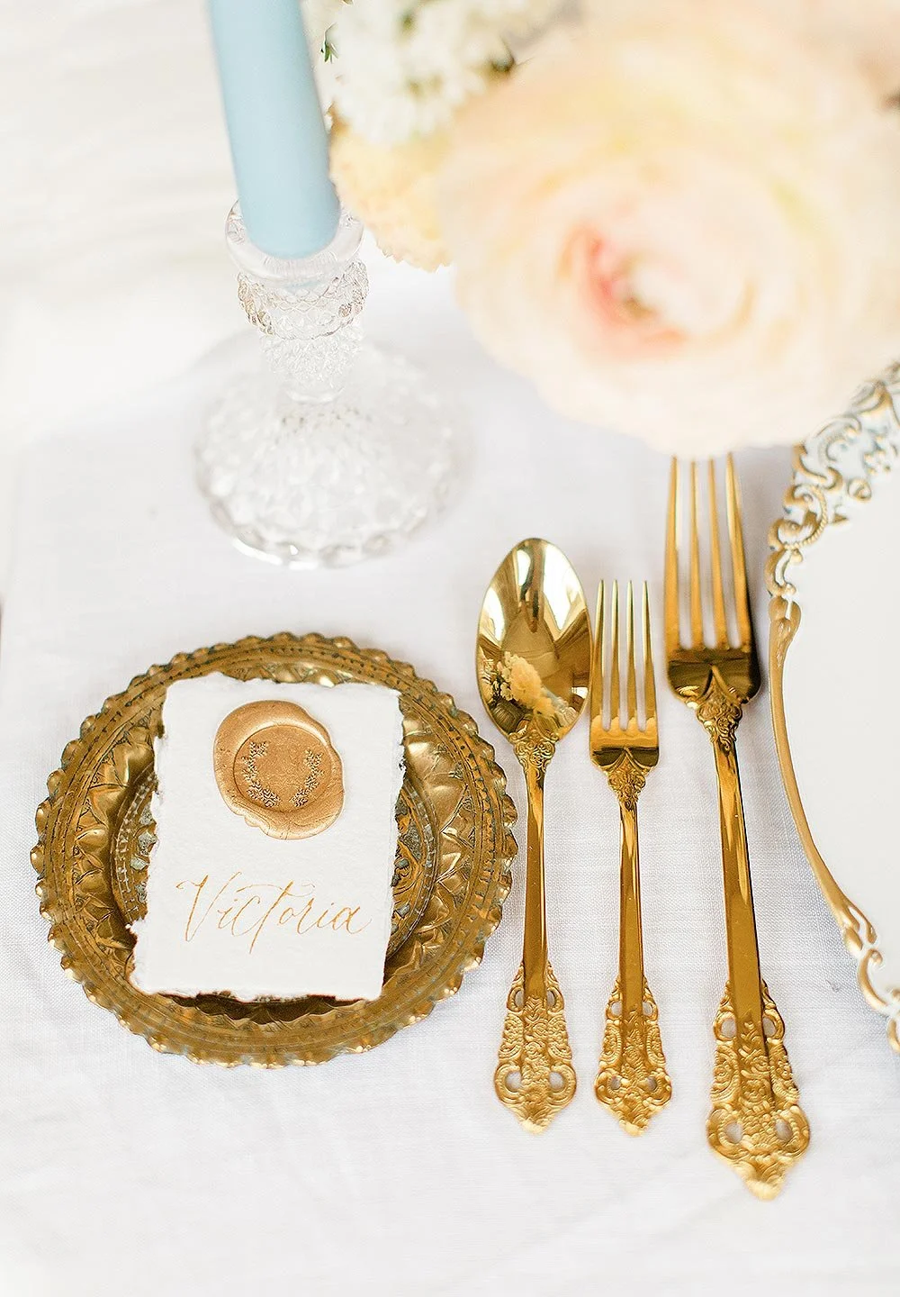 Elegant table setting with gold cutlery, a decorative gold charger plate, a white card with the name Victoria written in cursive, and a glass candle holder with a blue candle. There are light-colored flowers in the background.