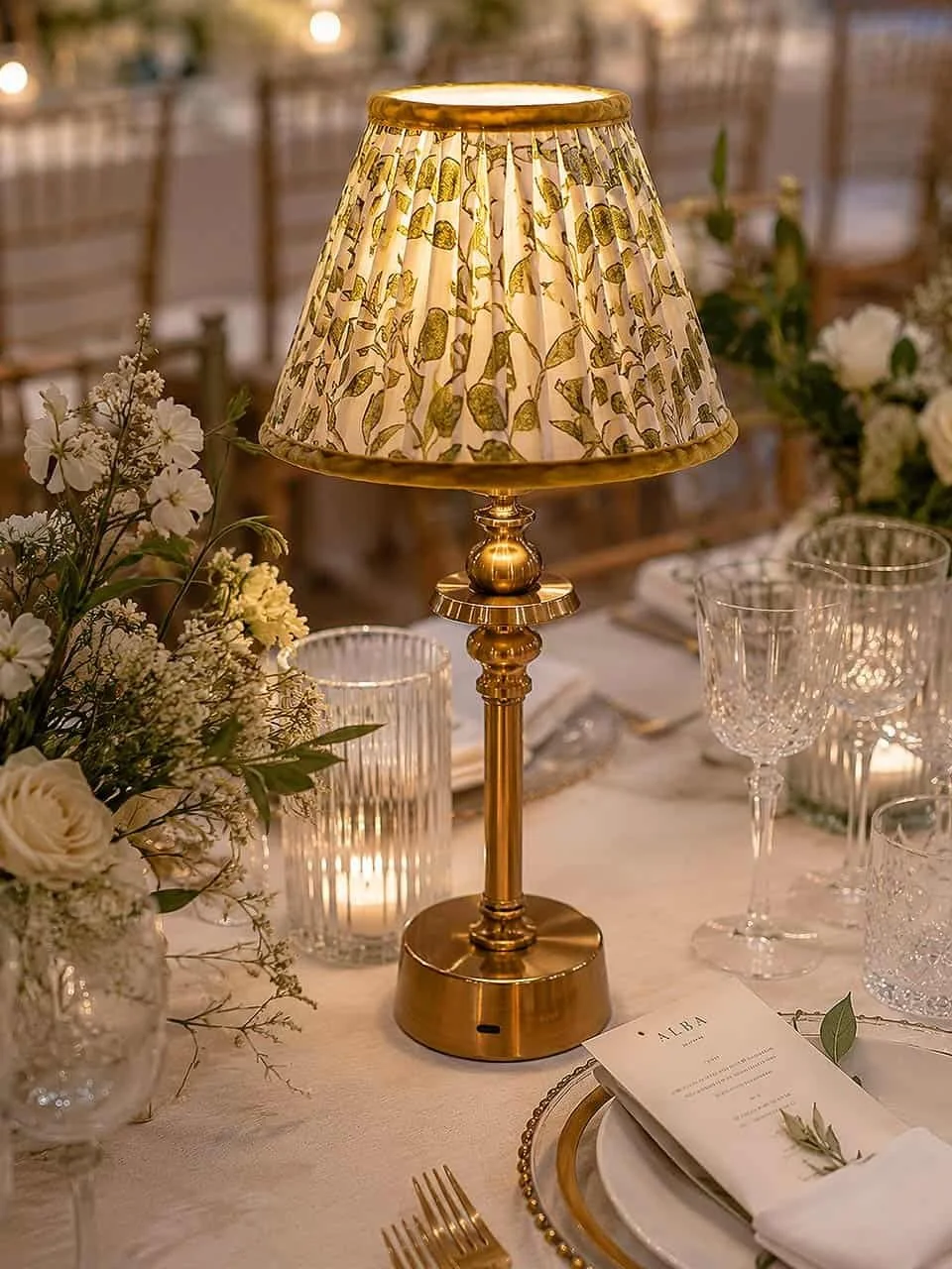 Elegant table setting with a brass lamp, floral centerpiece, candles, glassware, and a menu card.