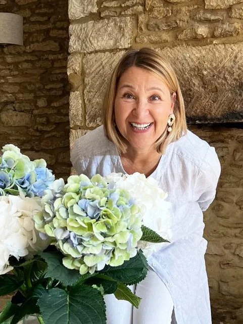 A woman smiling behind a bouquet of white and light blue hydrangeas, sitting in front of a stone fireplace.