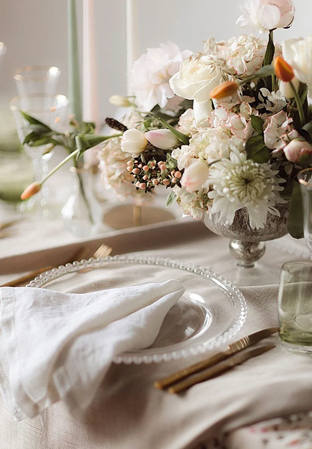 Elegant table setting with a large bouquet of white and blush pink flowers in a silver vase, a white napkin on a glass plate, gold-colored utensils, and a glass of water, set on a cream-colored tablecloth.