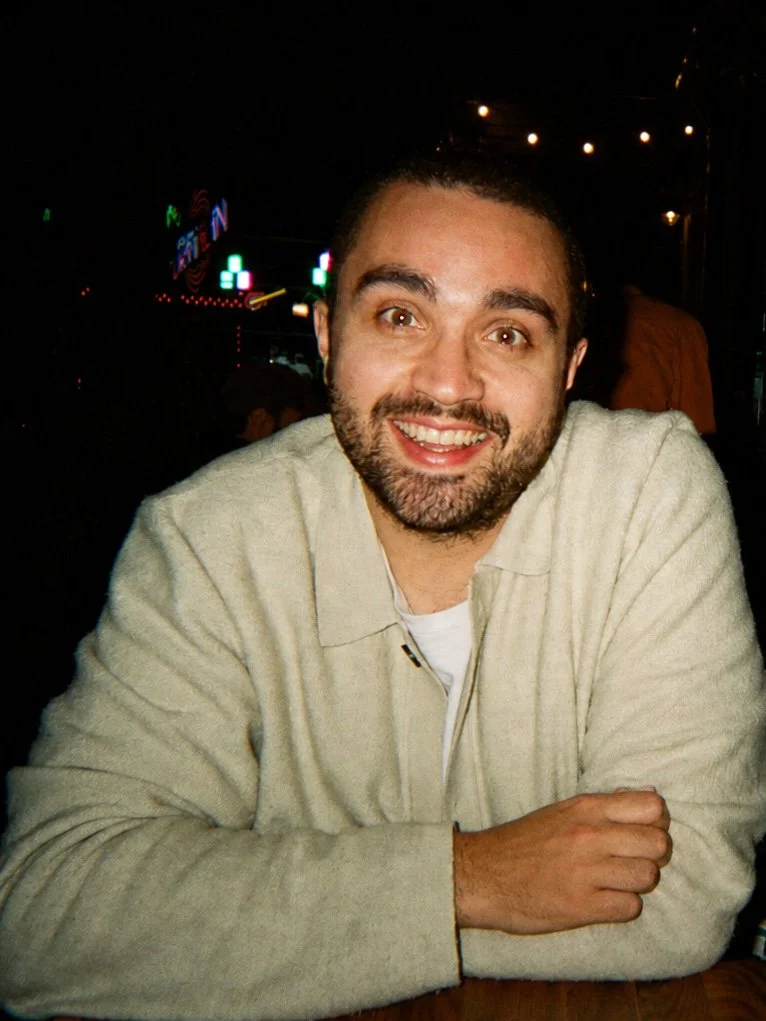 A smiling man with a beard and short dark hair, wearing a beige sweater, sitting at a table in a dimly lit setting, possibly a restaurant or bar.