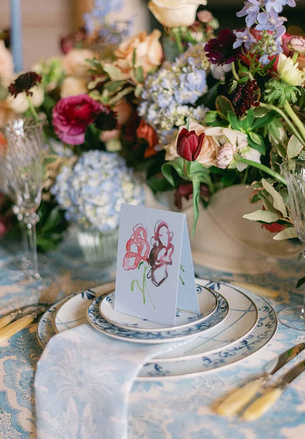 A table setting with blue and white patterned plates, a white napkin, and a card with an abstract watercolor flower design. The table is decorated with colorful flower arrangements in vases.