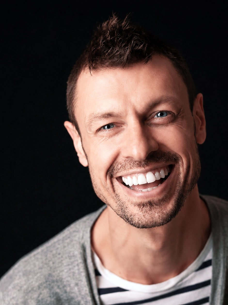 Close-up of a smiling man with blue eyes, short dark hair, and stubble, wearing a gray sweater over a striped shirt against a black background.