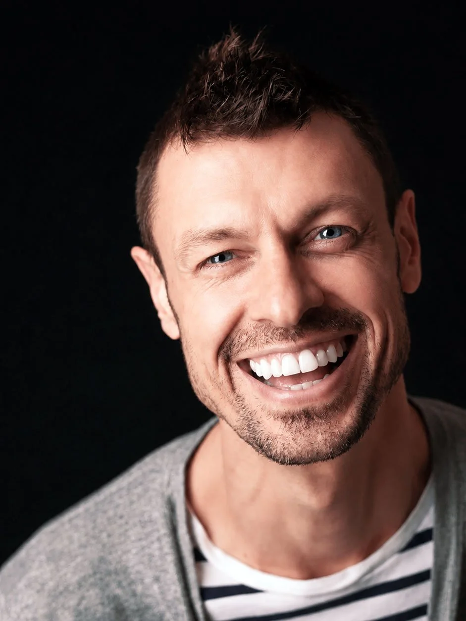 Close-up of a smiling man with blue eyes, short dark hair, and stubble, wearing a gray sweater over a striped shirt against a black background.