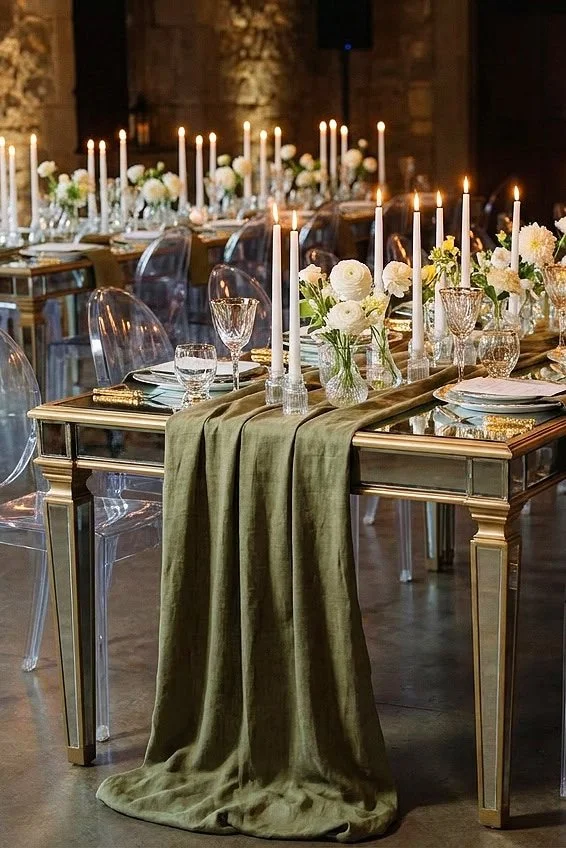 Elegant dining table set with white flowers, candles, glassware, and gold-accented tablecloths in a decorated event space.