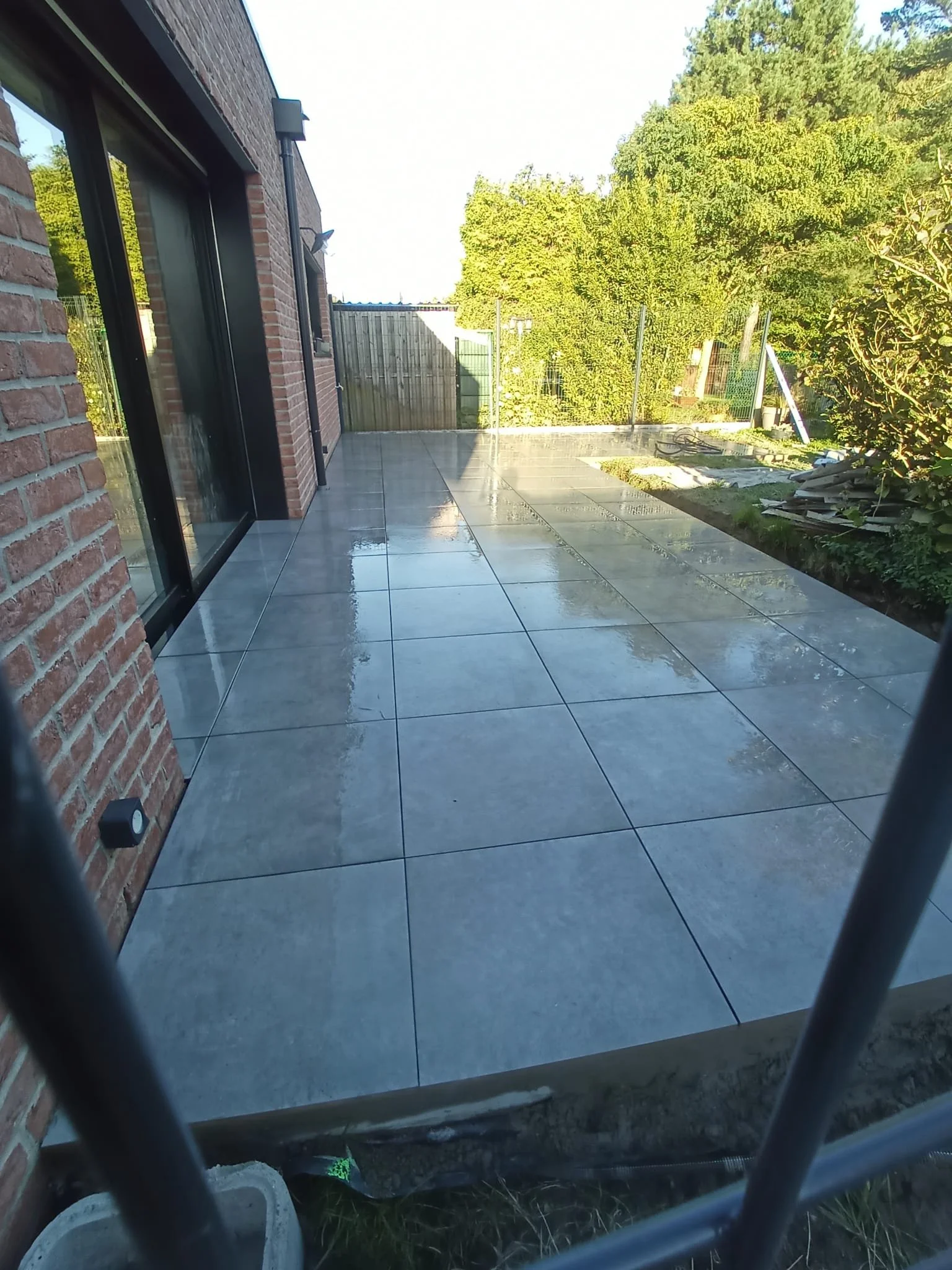 Terrasse en pierre réfléchissante avec un mur en briques, entourée d'arbres et de végétation, ensoleillée.