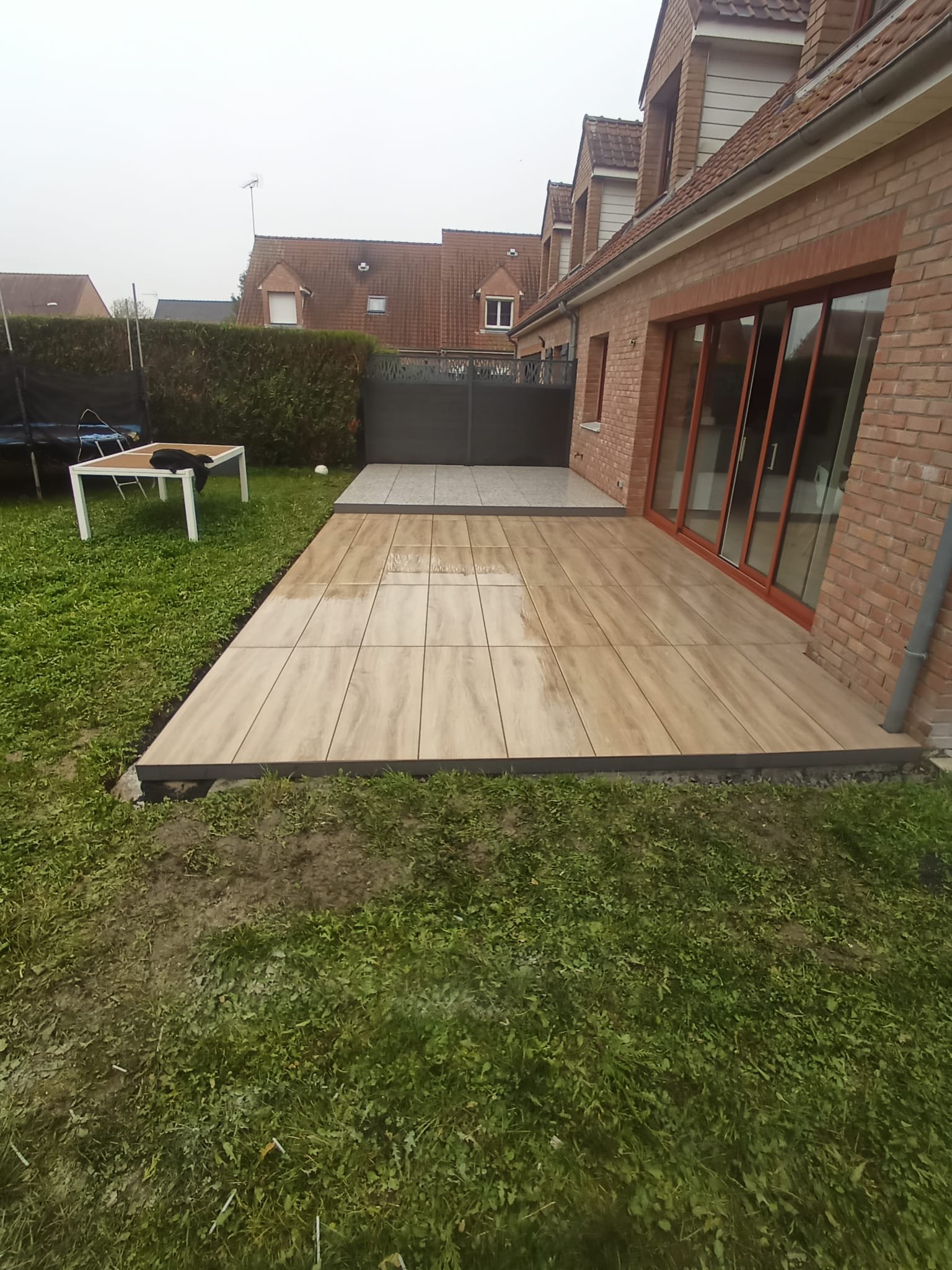 Une terrasse en bois adjacente à une maison en briques, avec une zone pavée, un jardin avec de l'herbe et un petit espace clôturé. Il fait gris et nuageux.