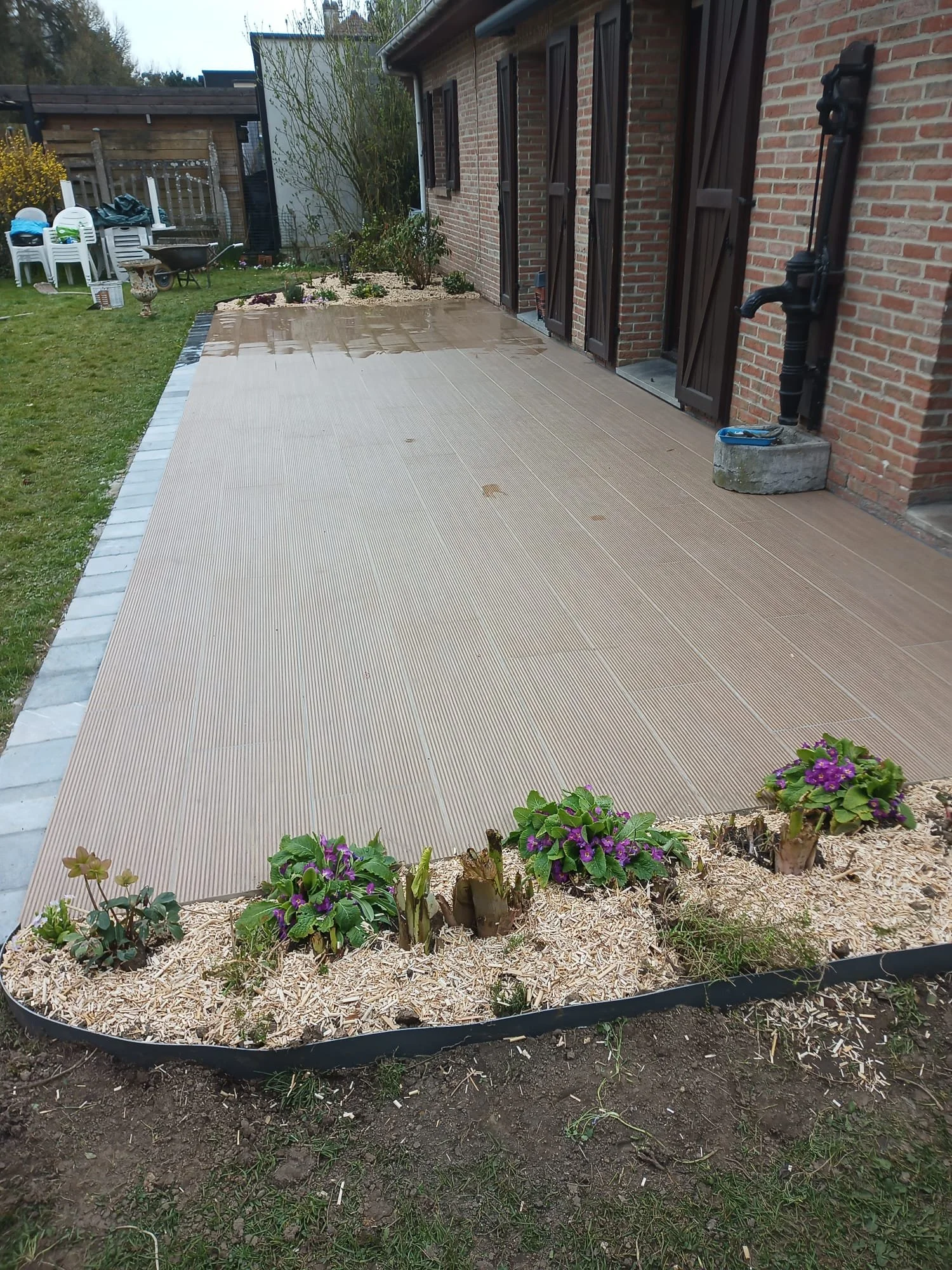 Un jardin avec une bordure en plastique noir, des plantes à fleurs violettes et vertes, une terrasse en bois, une maison en briques avec volets en bois marron, et des outils de jardinage