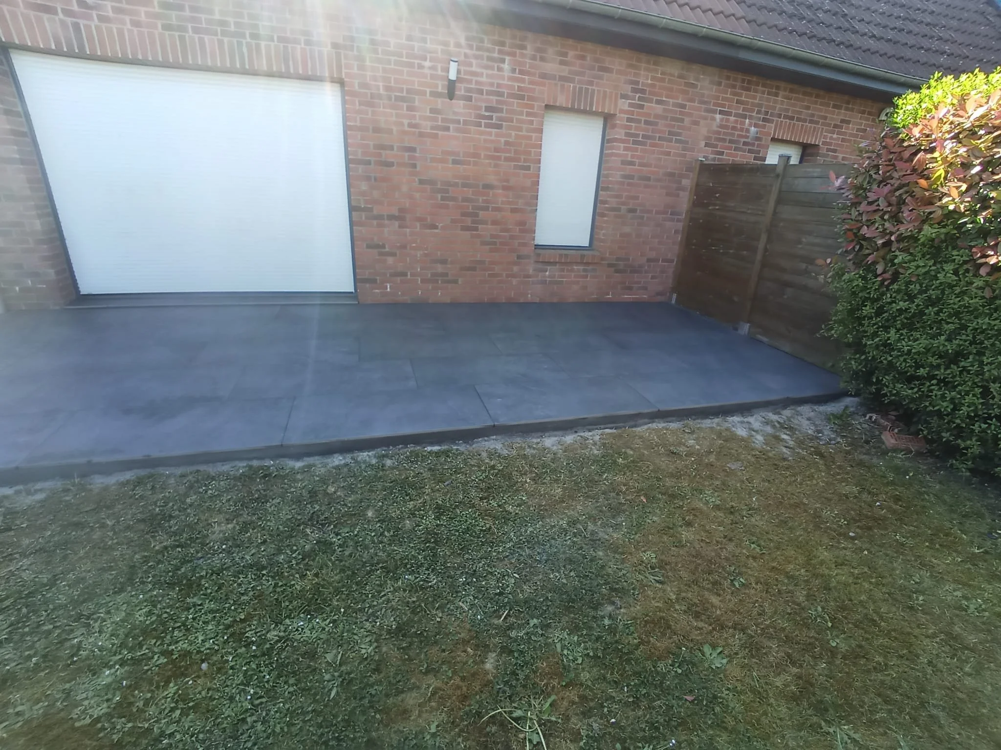 Une petite terrasse pavée devant une maison en brique avec un volet roulant blanc et un mur en bois, entourée de verdure.
