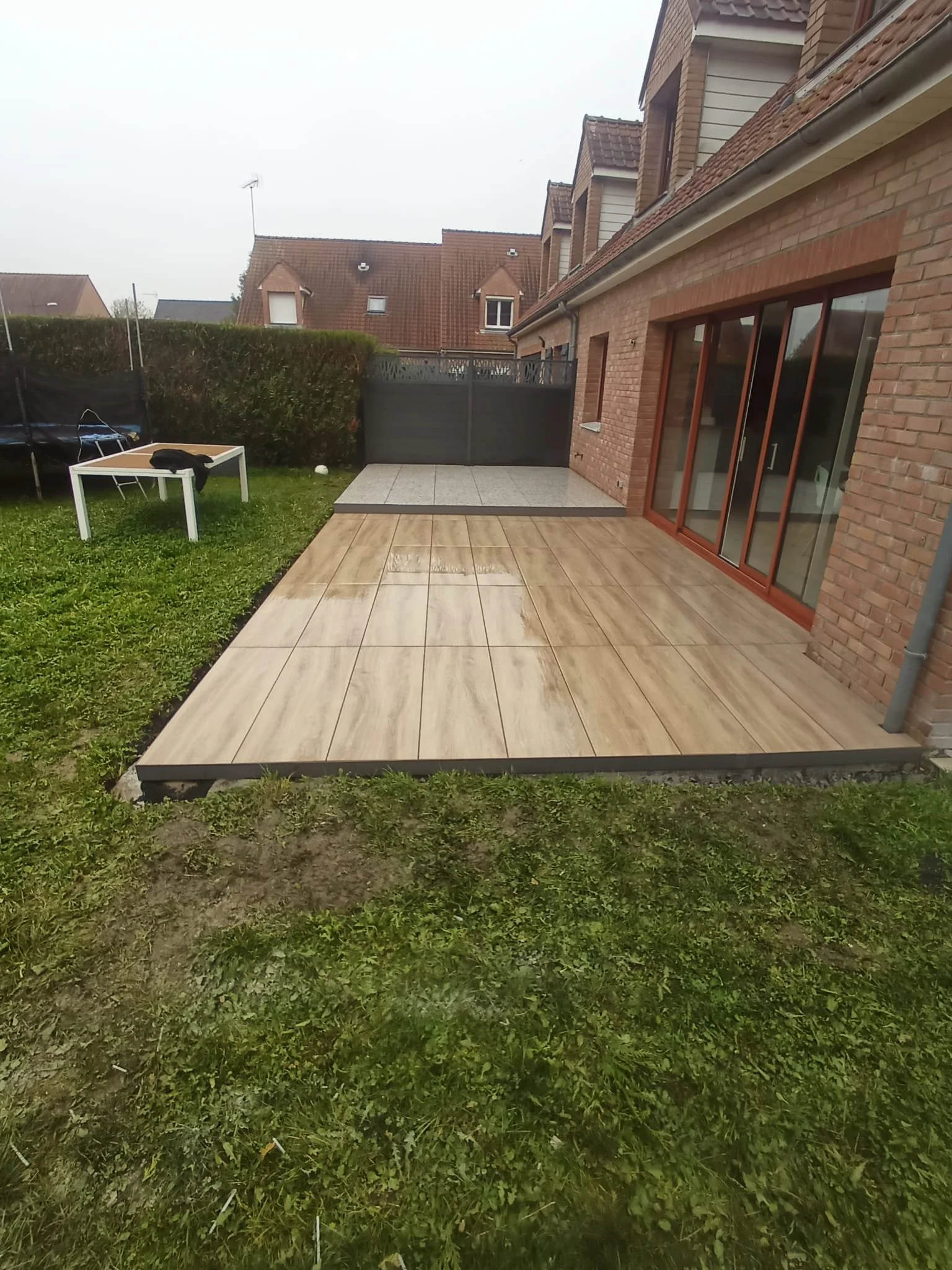  Terrasse en bois et carrelage devant une maison en briques avec grandes fenêtres en verre et porte-fenêtre.