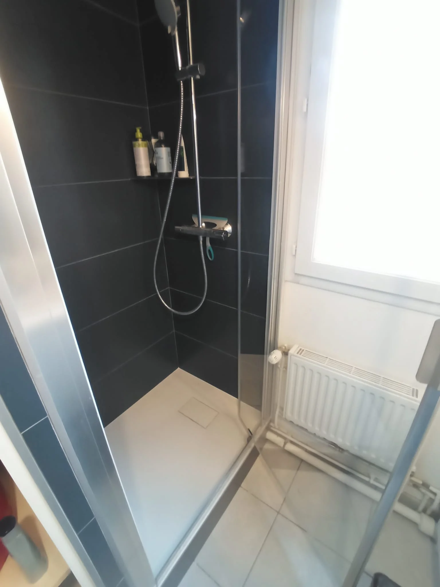 Douche avec murs en carreaux noirs, une rampe de douche, un porte-savon, une bouteille de shampooing, une fenêtre à droite, un radiateur en dessous, et un sol en carrelage blanc.