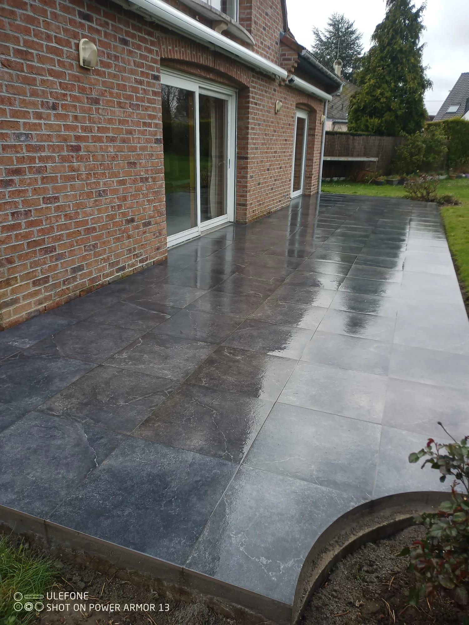 Une terrasse en pierre sombre, devant une maison en briques rouges avec deux portes-fenêtres en verre. La surface de la terrasse est humide, reflétant la lumière. Un coin de jardin avec des plantes en pot est visible dans le coin inférieur droit.