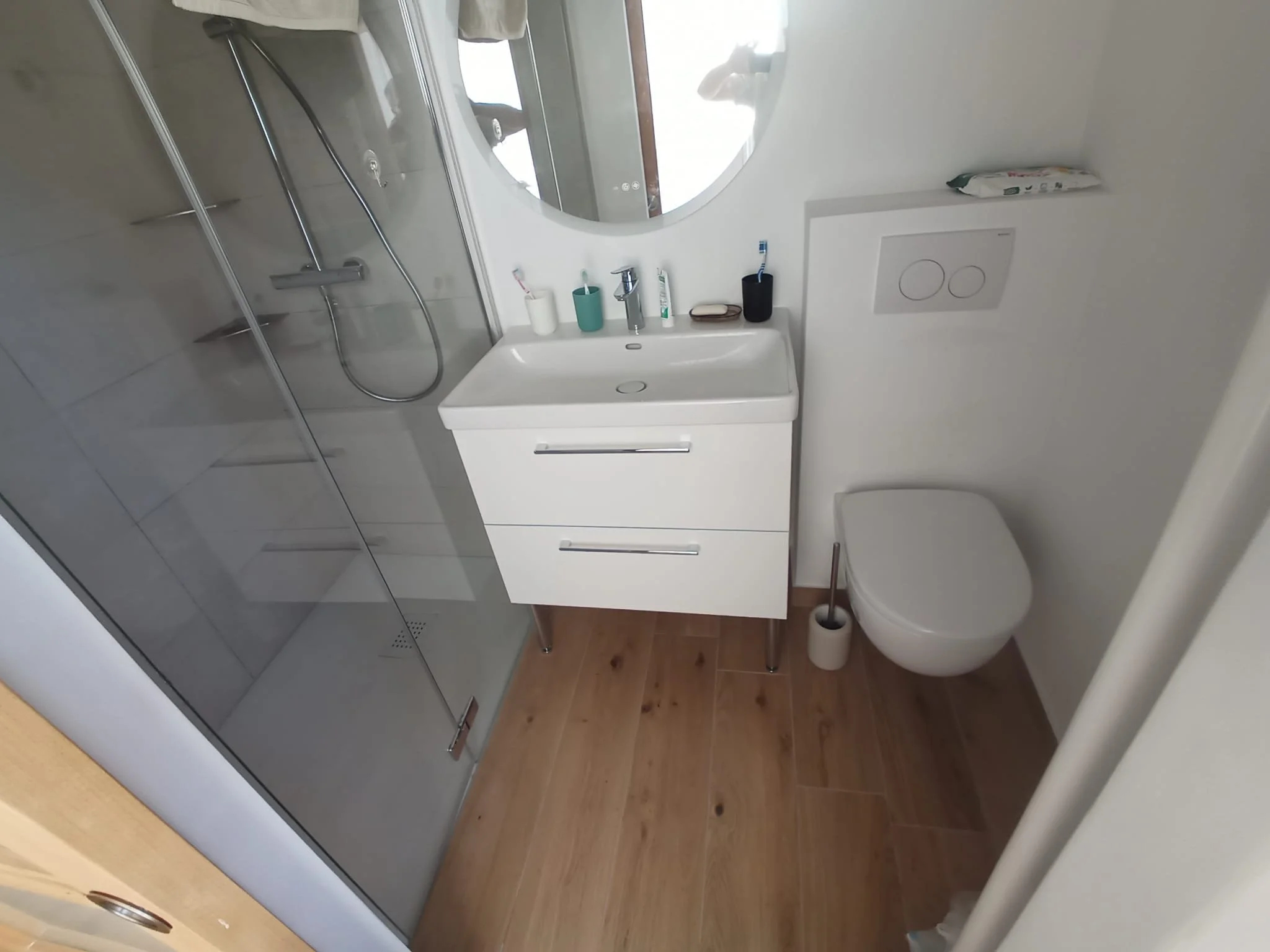 Petite salle de bain moderne avec douche, miroir rond, lavabo blanc avec rangement, accessoires de toilette, toilettes suspendues et brosse WC, sol en bois clair.