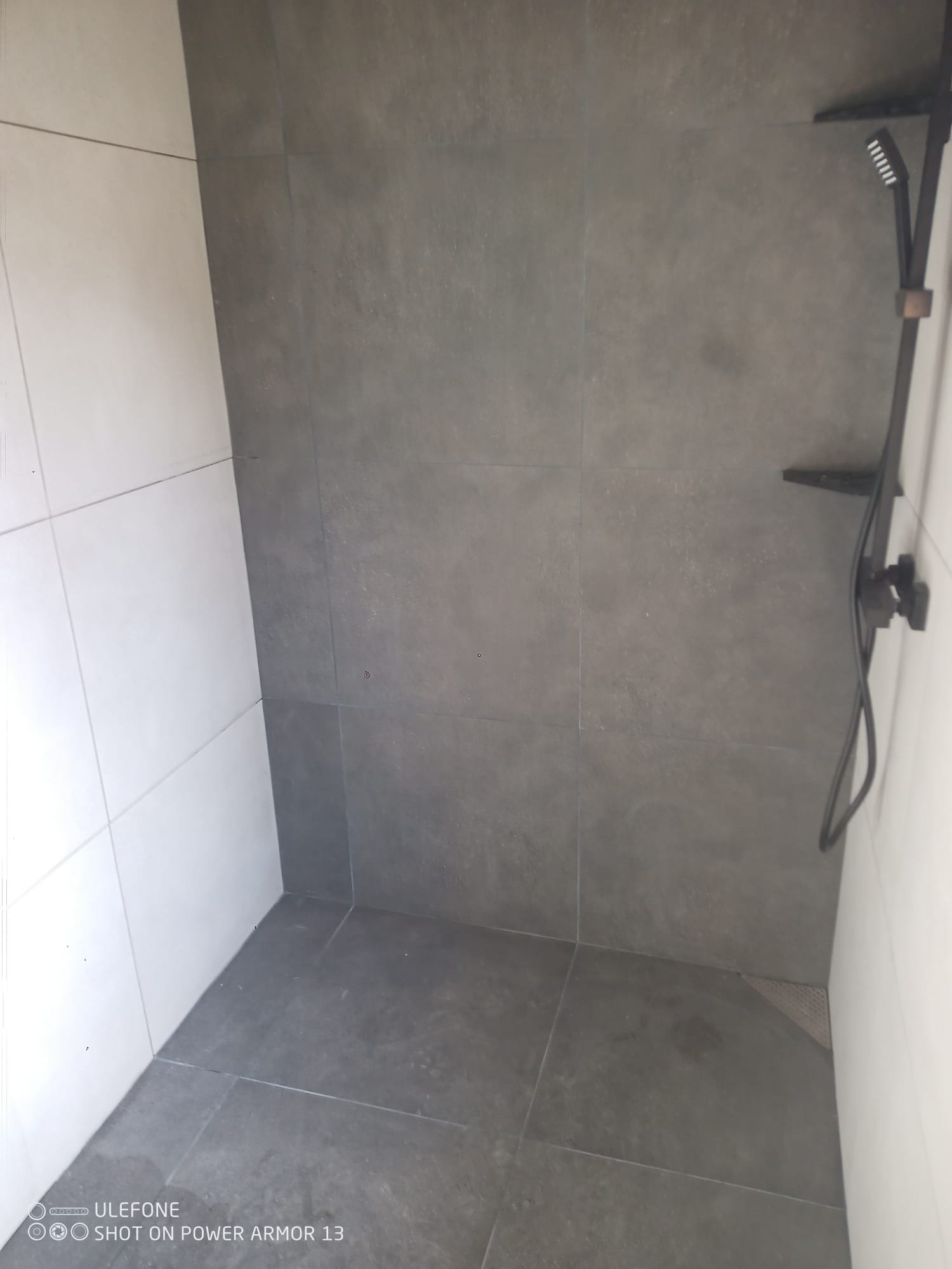 Salle de bain avec des murs et le sol en carrelage gris foncé, une douche ouverte sans porte, avec une barre de maintien. La photo est prise de côté, montrant principalement un mur et le sol.
