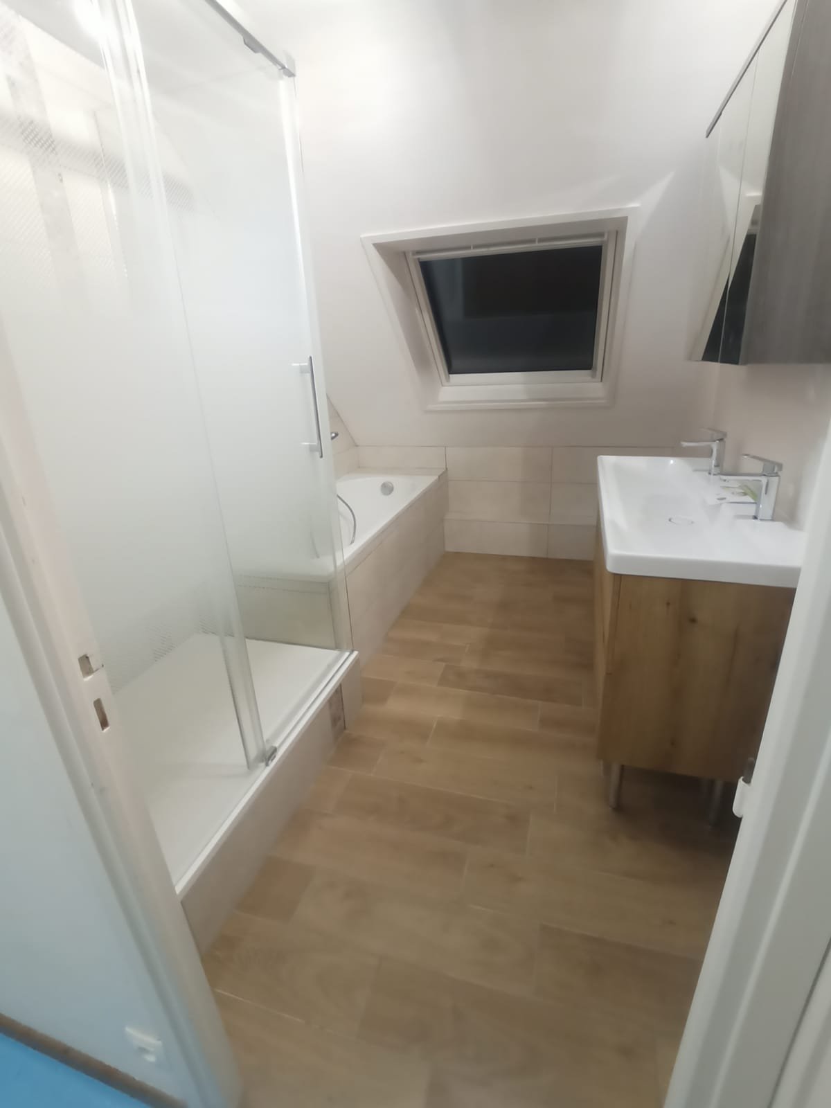 Petite salle de bain moderne avec douche, baignoire, lavabo avec meuble en bois, fenêtre de toit et sol en bois.