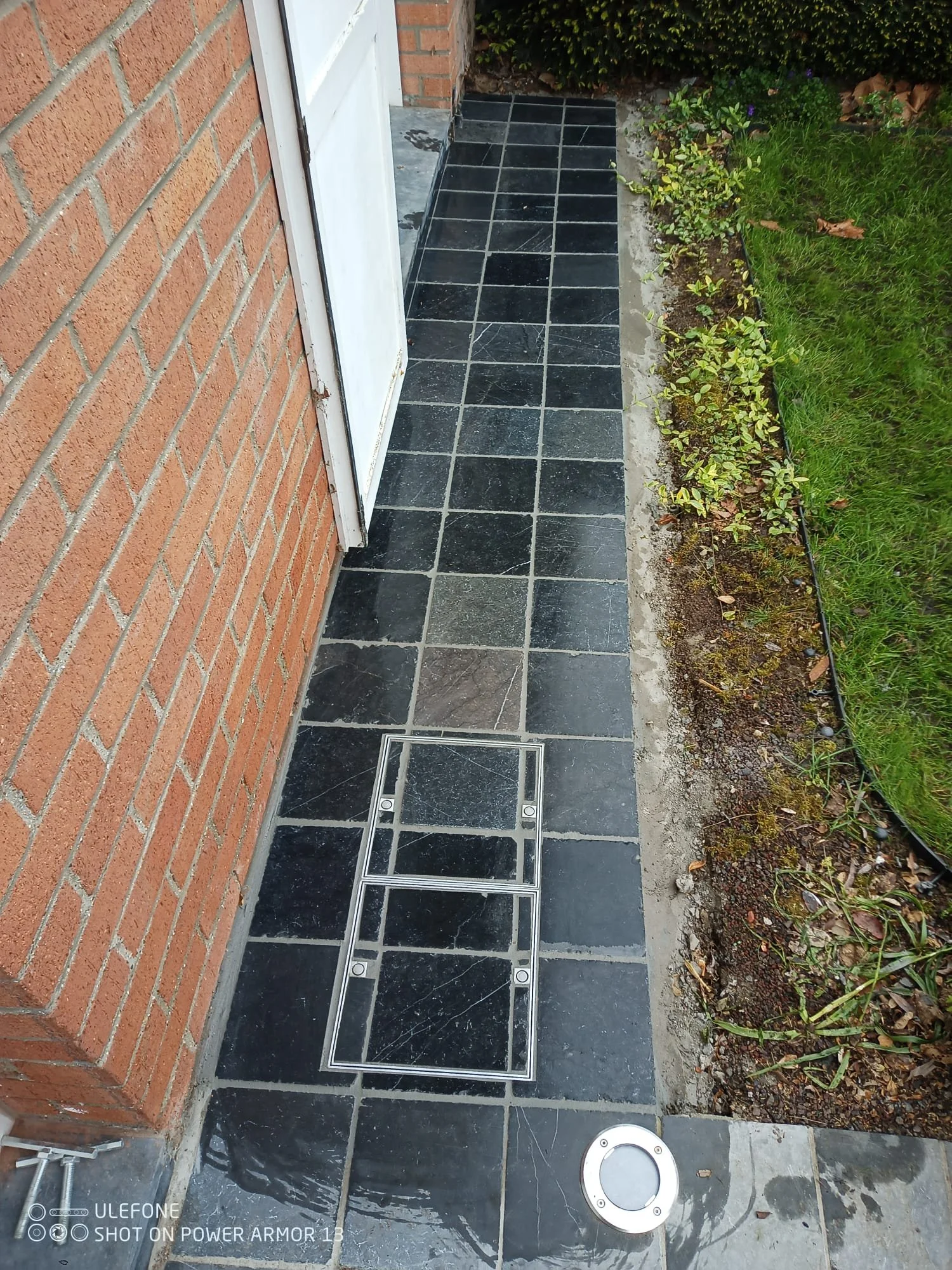 Entrée de maison avec sol en carreaux noirs, une grille d'accès à une installation souterraine, un éclairage encastré, et un jardin avec des plantes à droite.