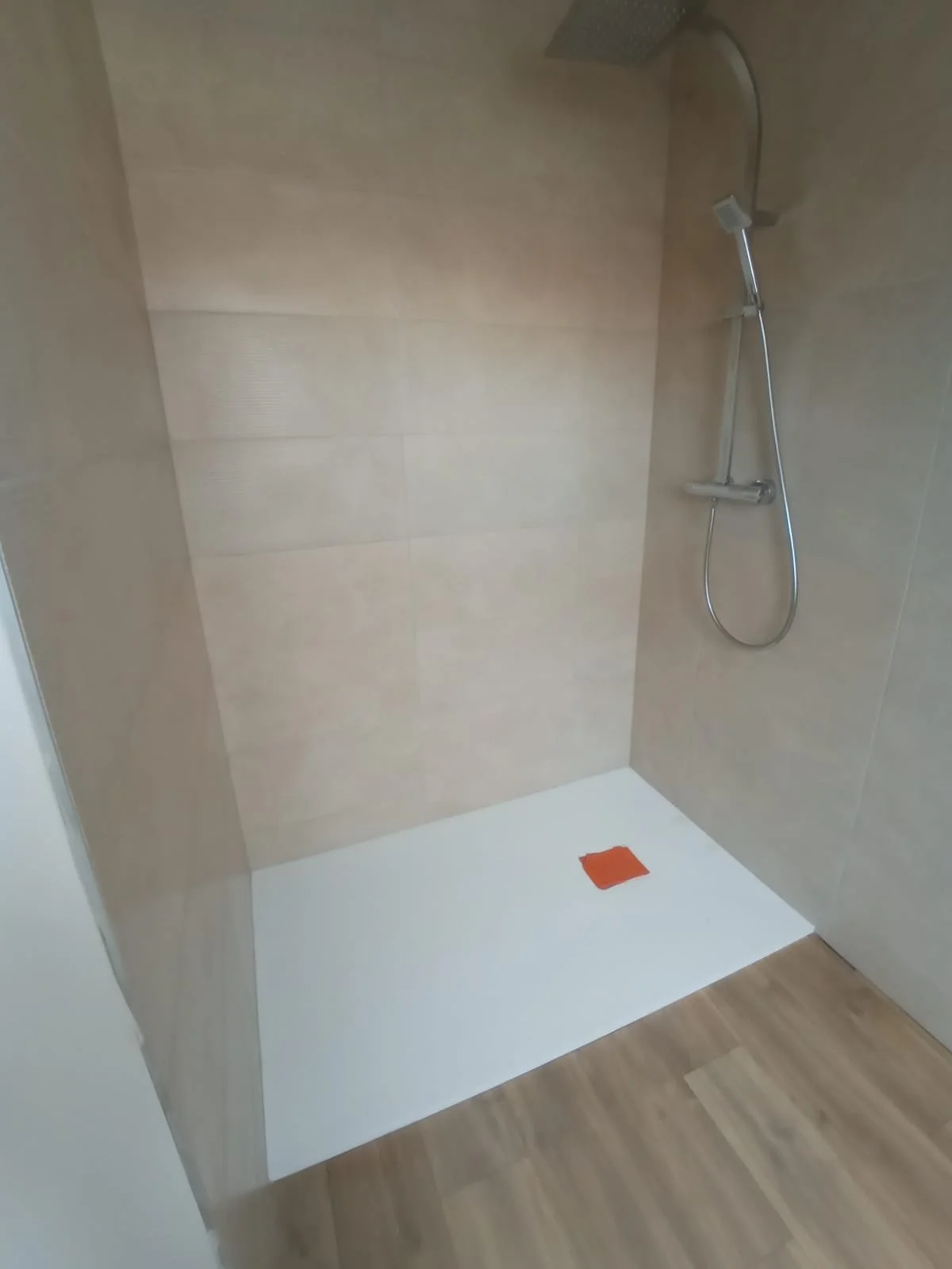 Douche neuve avec un revêtement en carrelage beige, une pomme de douche fixe et une douchette amovible, sol en bois, avec une trappe de sol orange.