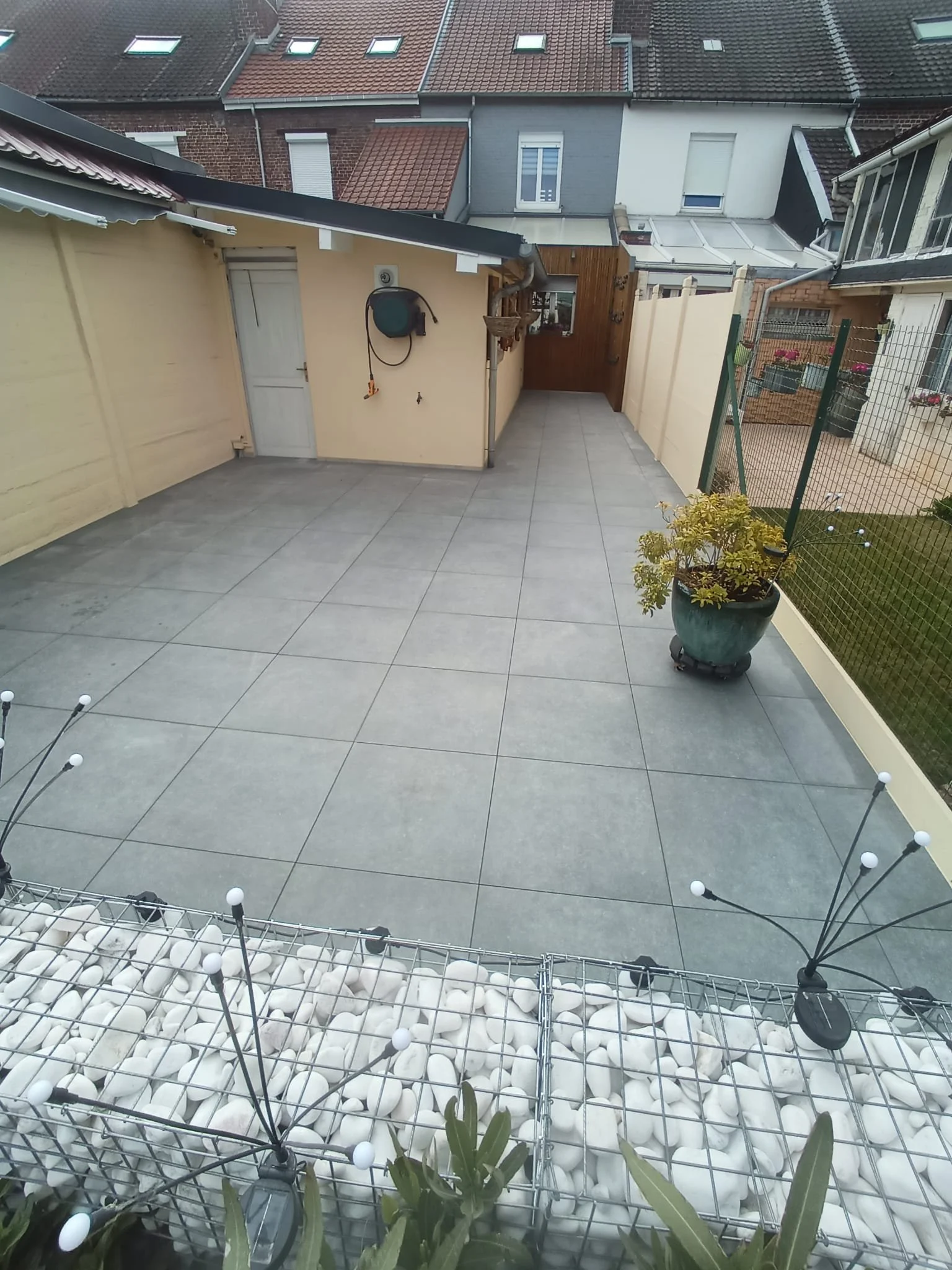 Terrasse urbaine avec sol en carrelage gris, entourée de murs en plâtre beige et clôture en métal. Il y a une grande plante en pot, un petit stand en métal avec des pierres, et un petit bâtiment avec un tuyau d'arrosage. En arrière-plan, on voit des 