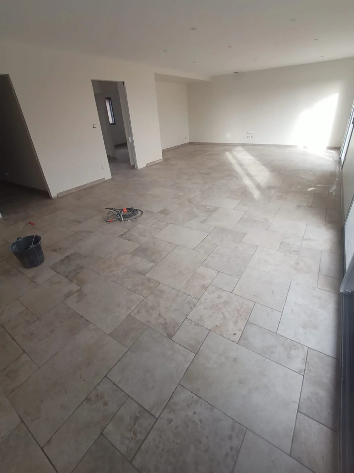 Pièce de salon avec sol en carrelage beige, murs blancs, et fenêtres laissant entrer la lumière naturelle, en cours de rénovation.