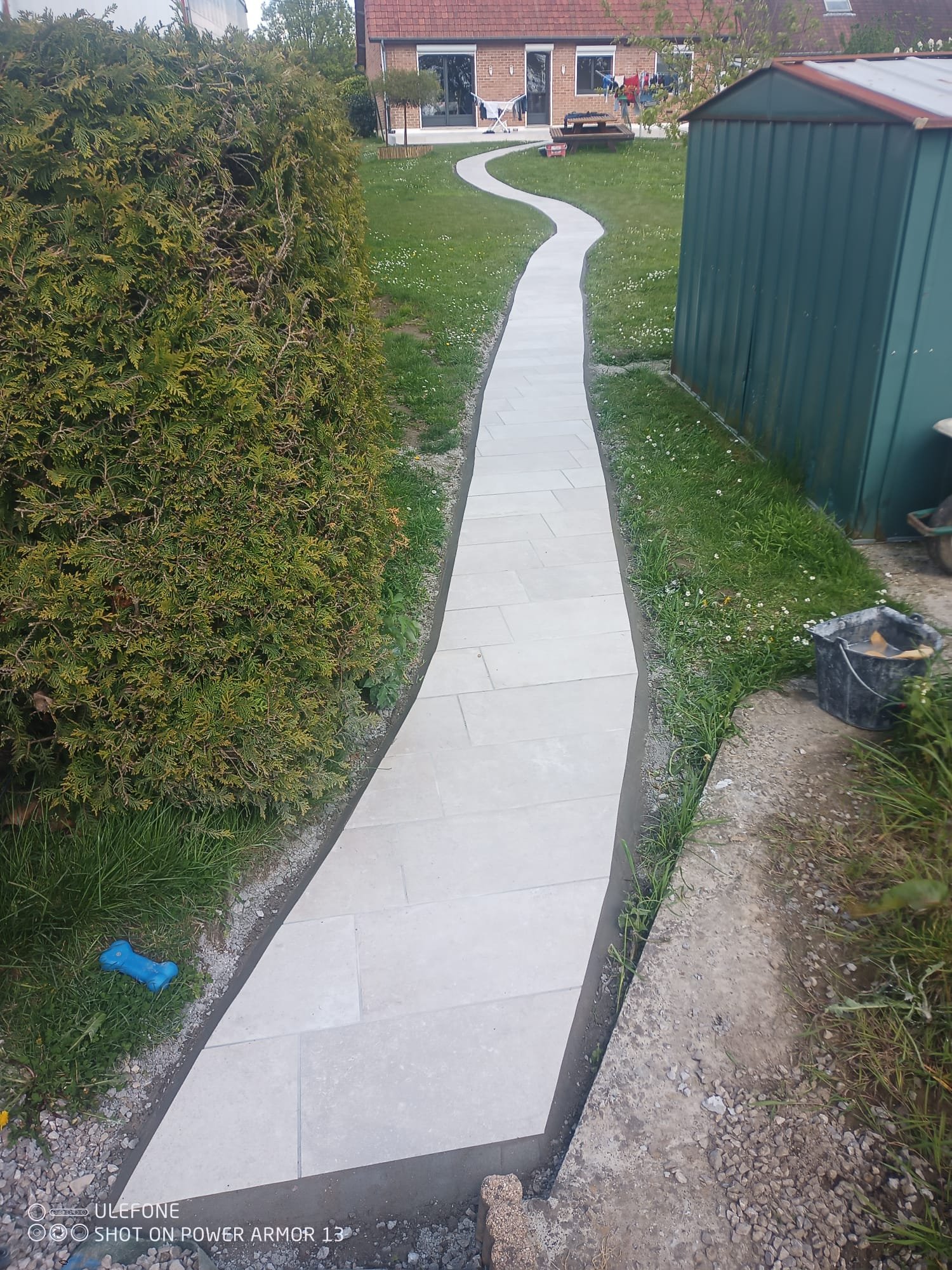 Chemin en pierre qui serpente dans un jardin au devant d'une maison avec une terrasse et des vêtements suspendus à une corde à linge.