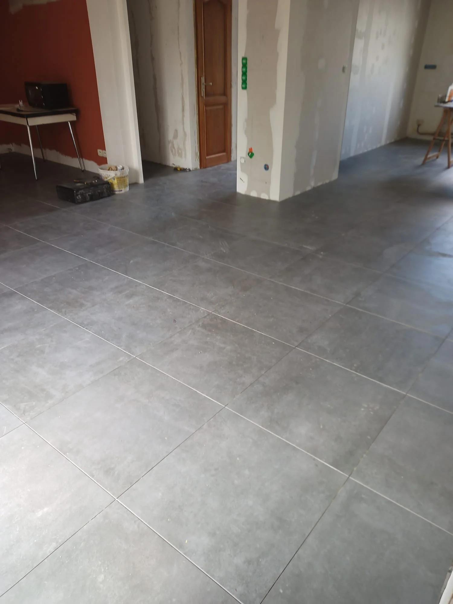 Une pièce en rénovation avec des murs partiellement peints et des carreaux gris au sol. Il y a un petit bureau avec une radio et des outils dessus, ainsi qu'un seau de peinture. Un mur porte des prises électriques et des interrupteurs, et il y a un tabouret pliant dans un coin.