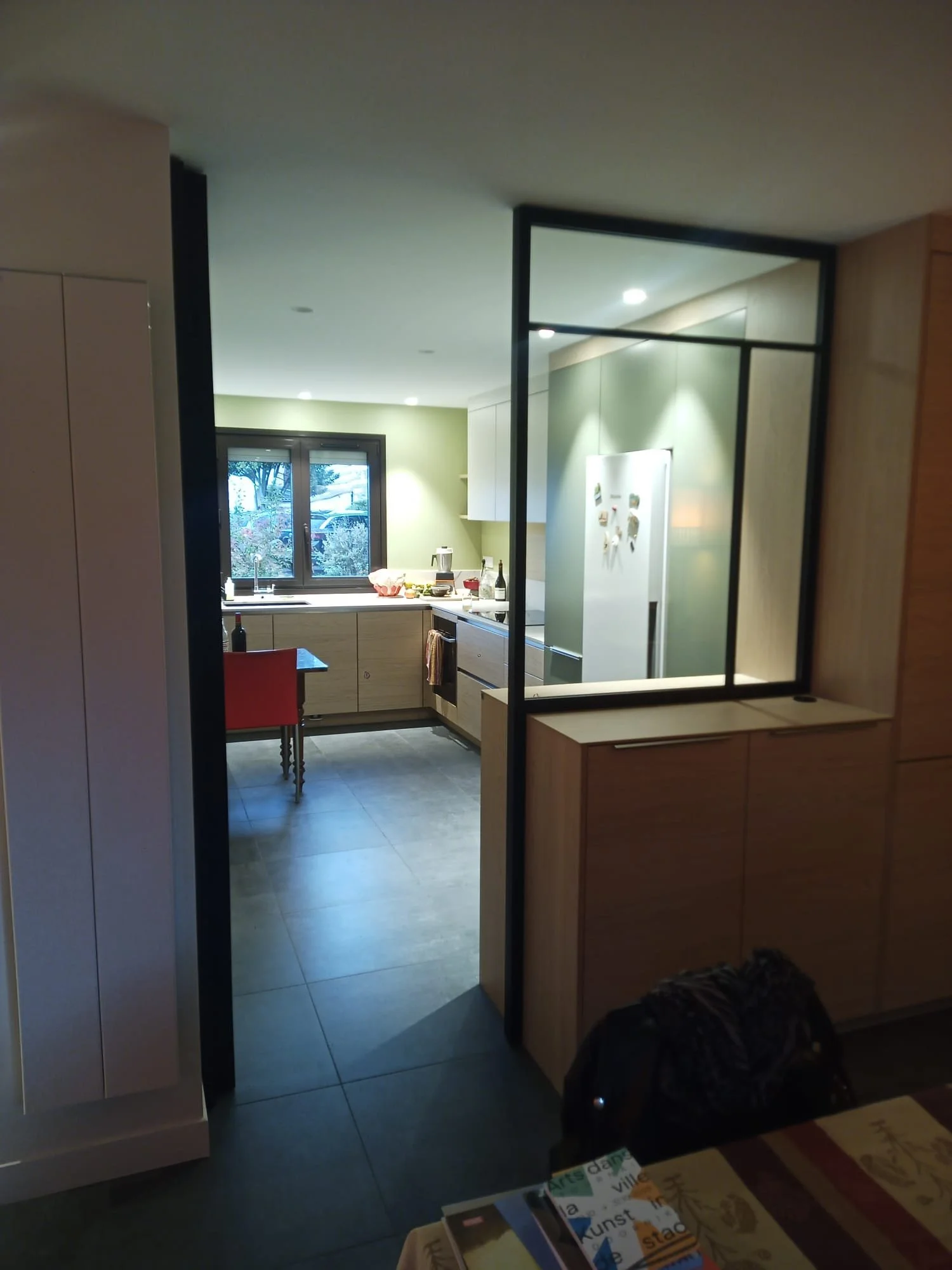 Cuisine ouverte avec fenêtre, réfrigérateur blanc, meubles en bois clair, plan de travail, table rouge et chaise, vue vers l'extérieur de la maison.
