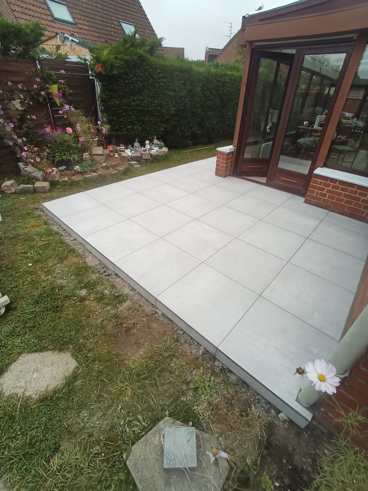 Terrasse en carrelage gris devant une maison avec des fenêtres en verre et un volet en bois, entourée de quelques végétaux et une petite zone de jardin avec des fleurs.